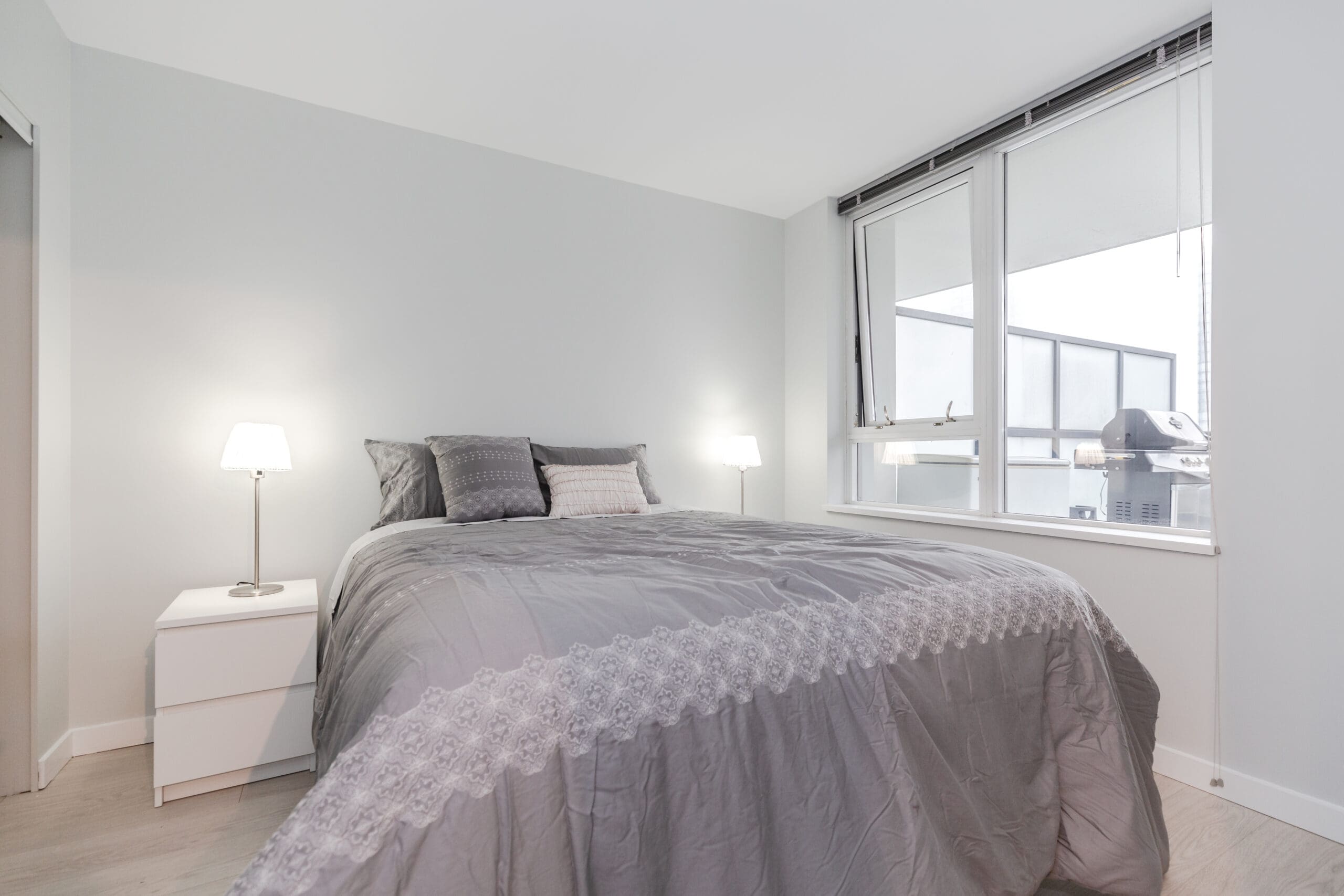 Minimalist bedroom with a gray duvet on a double bed, two bedside lamps, and a window showing a balcony with a grill.