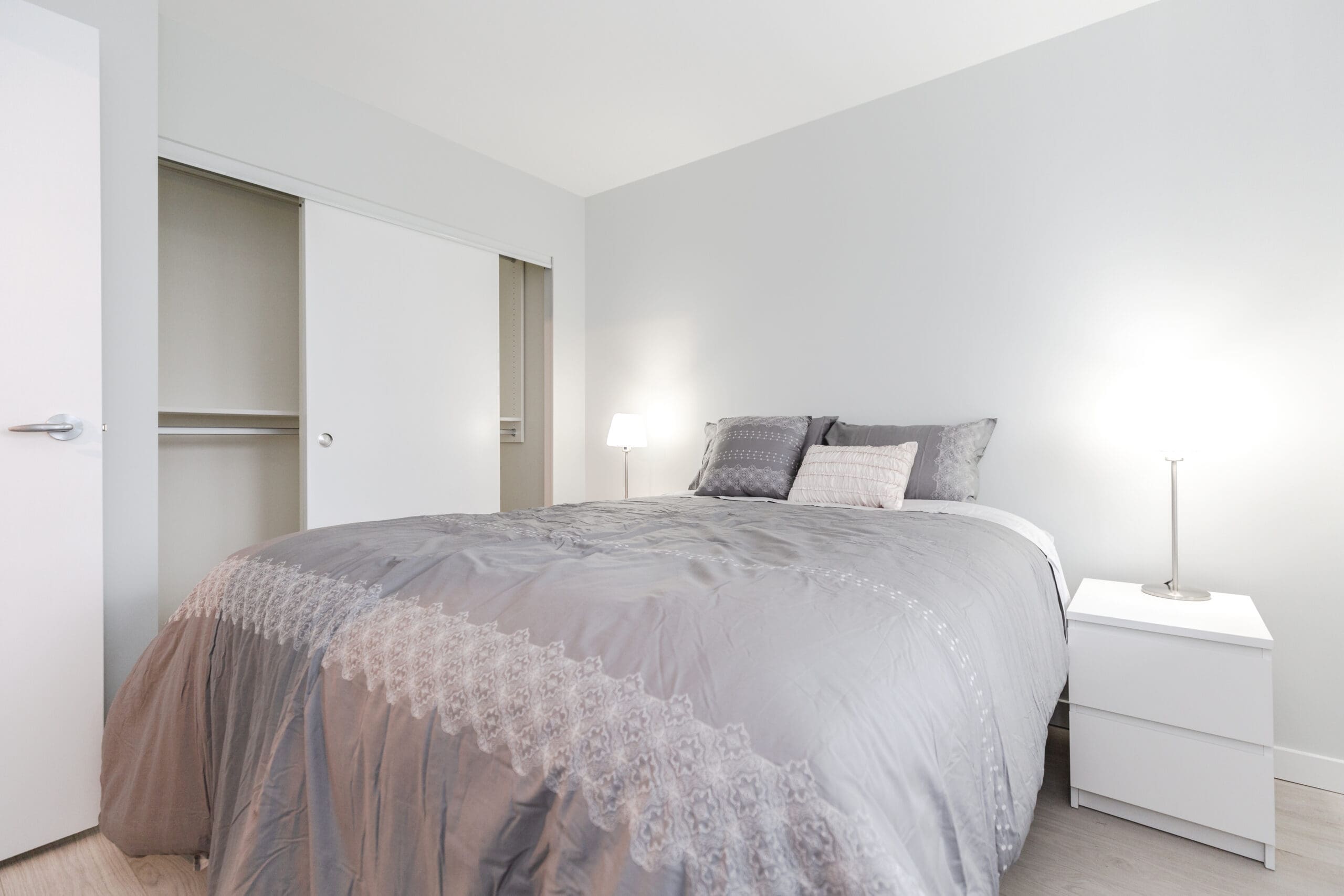 A neatly made bed with gray bedding in a minimalistic bedroom. A nightstand with a lamp is beside the bed, and an open sliding door closet is visible.