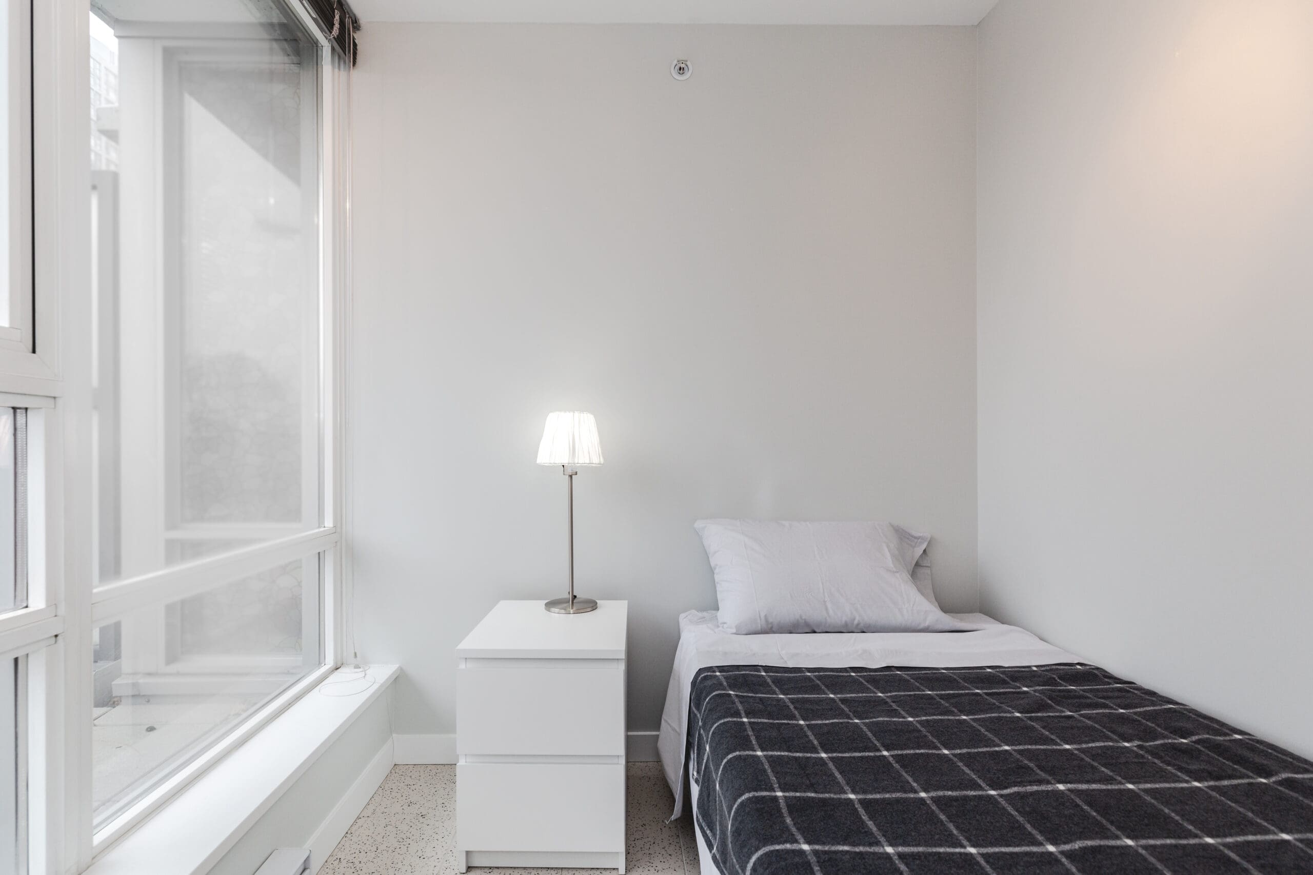 A minimalist bedroom with a single bed, a checked blanket, a white pillow, a bedside table, and a lamp by a large window with sheer curtains.