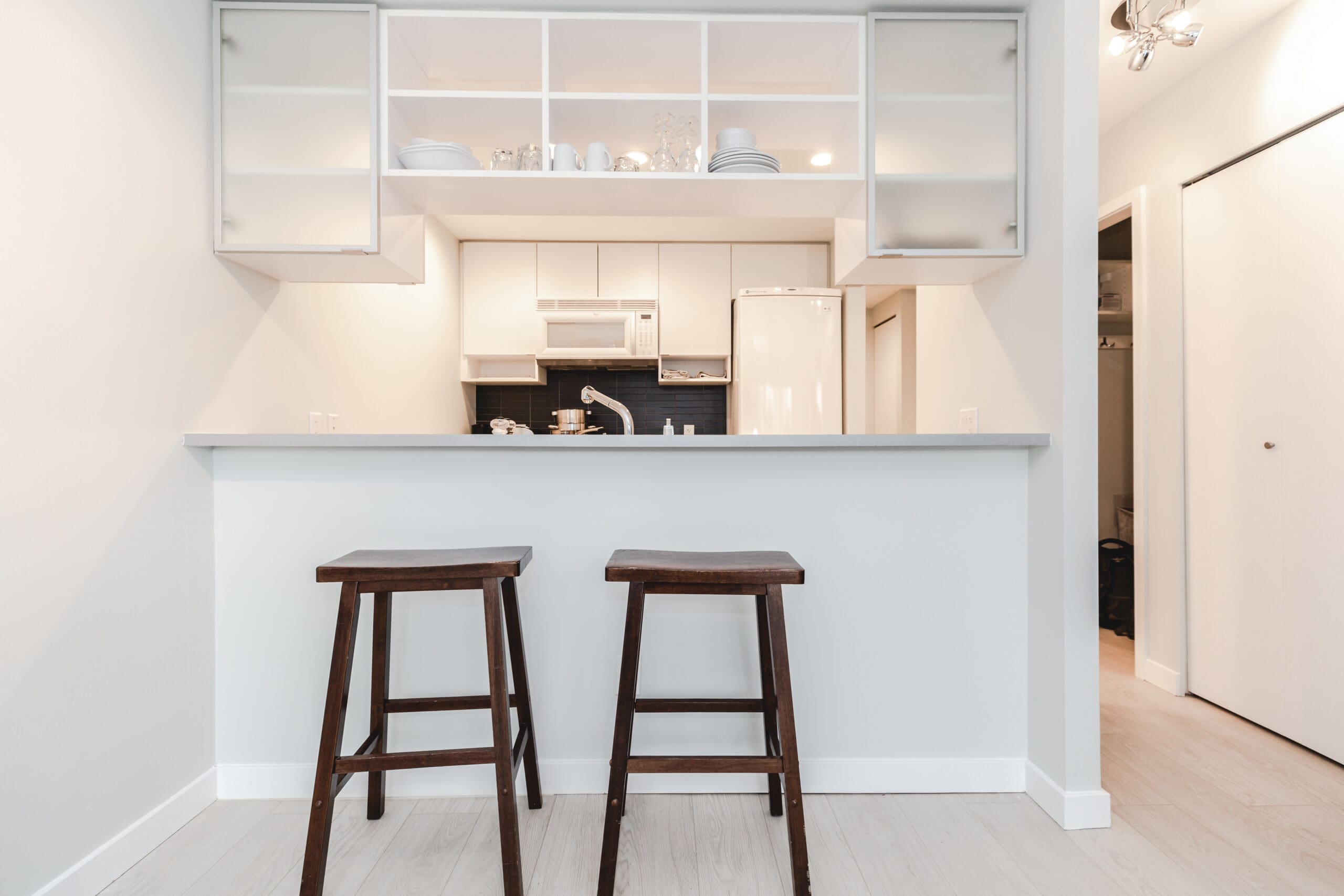 A minimalist kitchen with two wooden bar stools at a counter. Cabinets and appliances are in the background. The decor is neutral and modern.