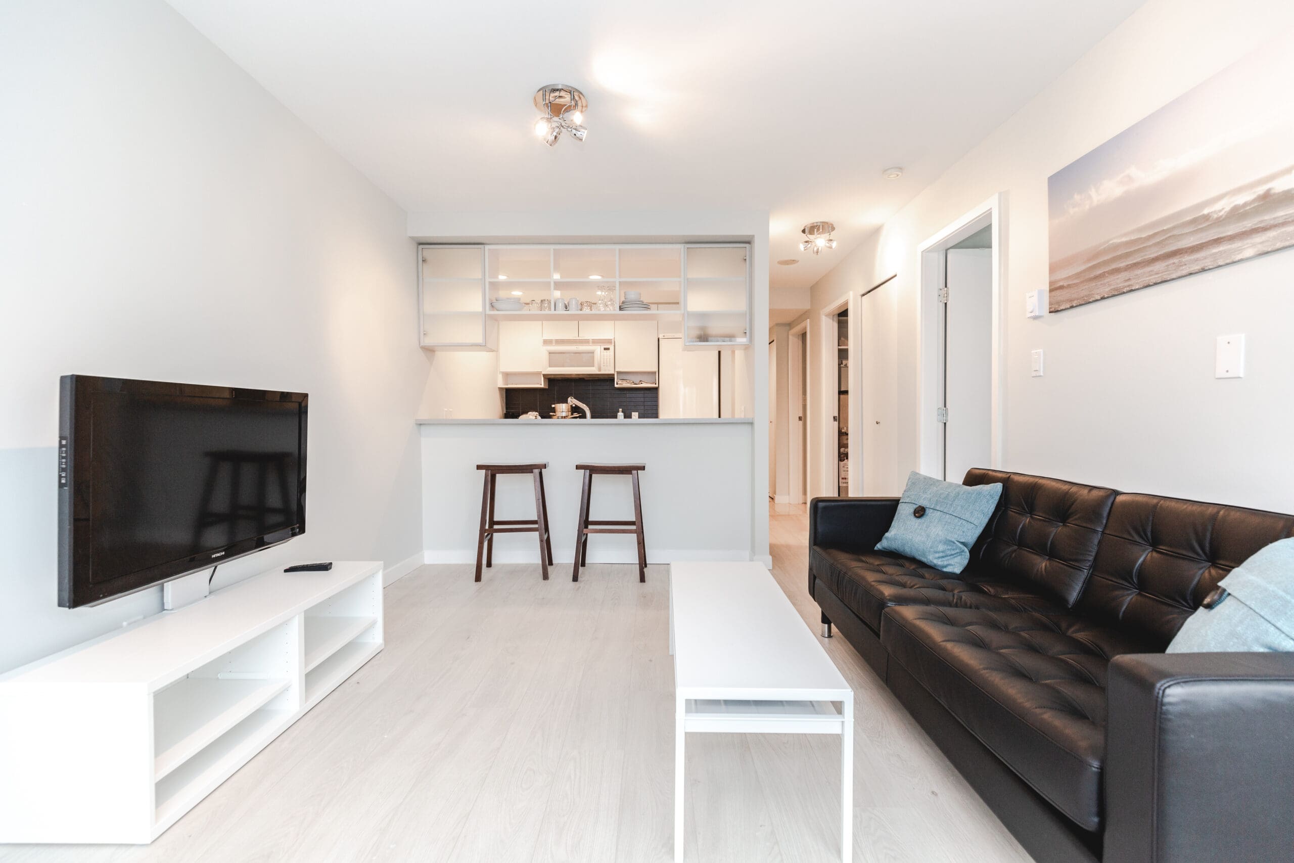 Modern living room with a black sofa, white coffee table, and TV on a stand. There's a kitchen bar with two stools and a framed picture on the wall. White walls and light wood flooring.