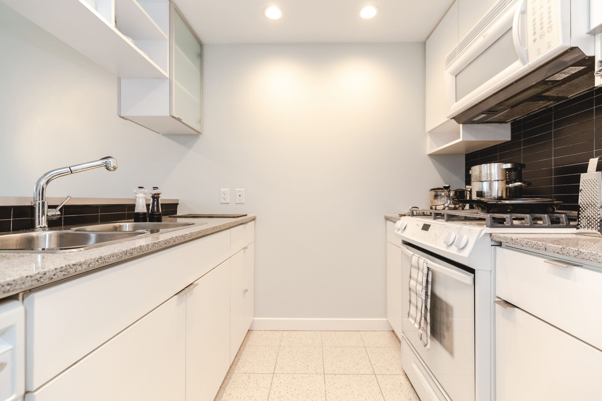 Modern kitchen with white cabinetry, stainless steel sink, black tile backsplash, and a gas stove. A striped towel hangs on the oven handle.
