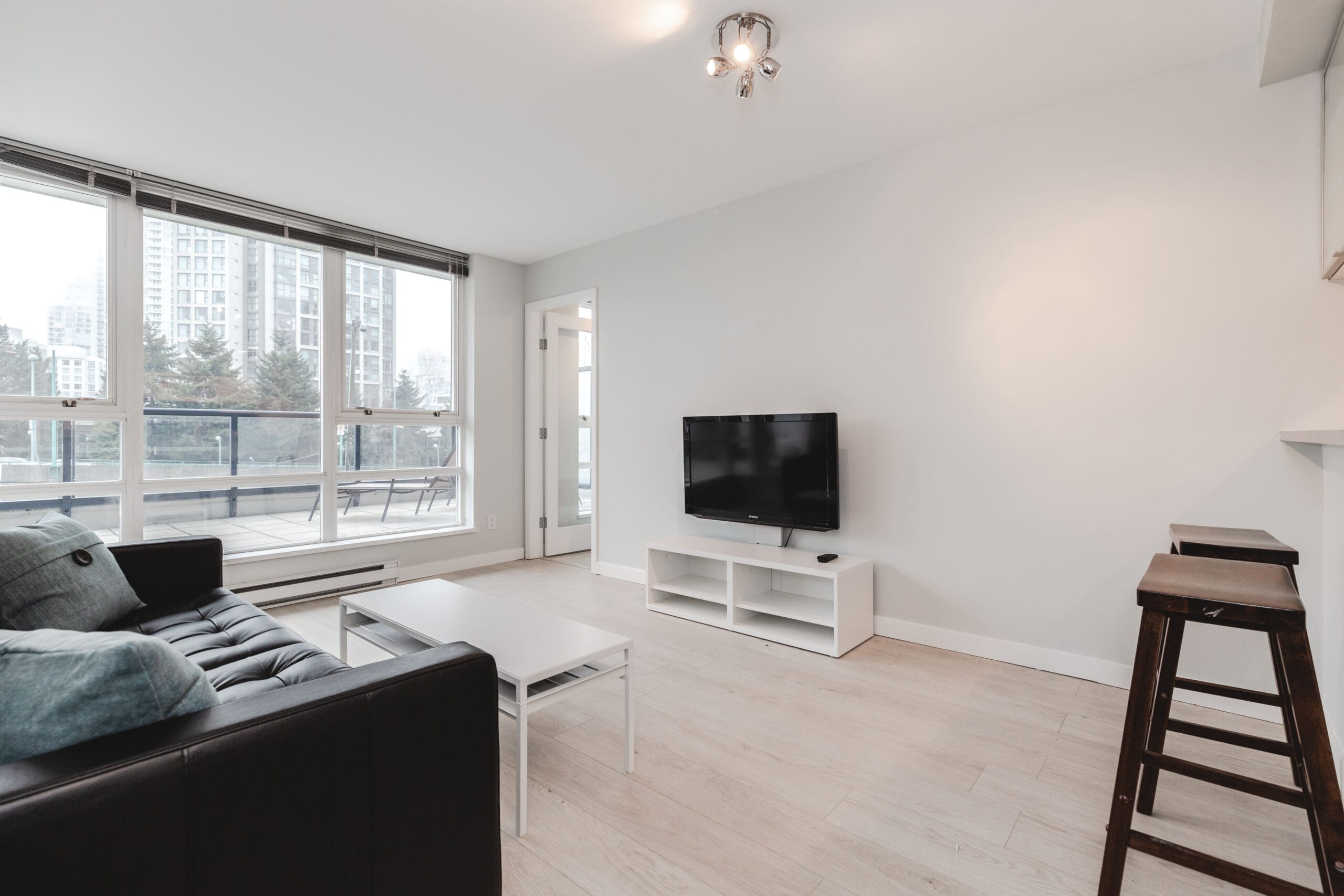Minimalist living room with a black sofa, white coffee table, wall-mounted TV on a stand, and two bar stools. Large windows offer a view of city buildings.