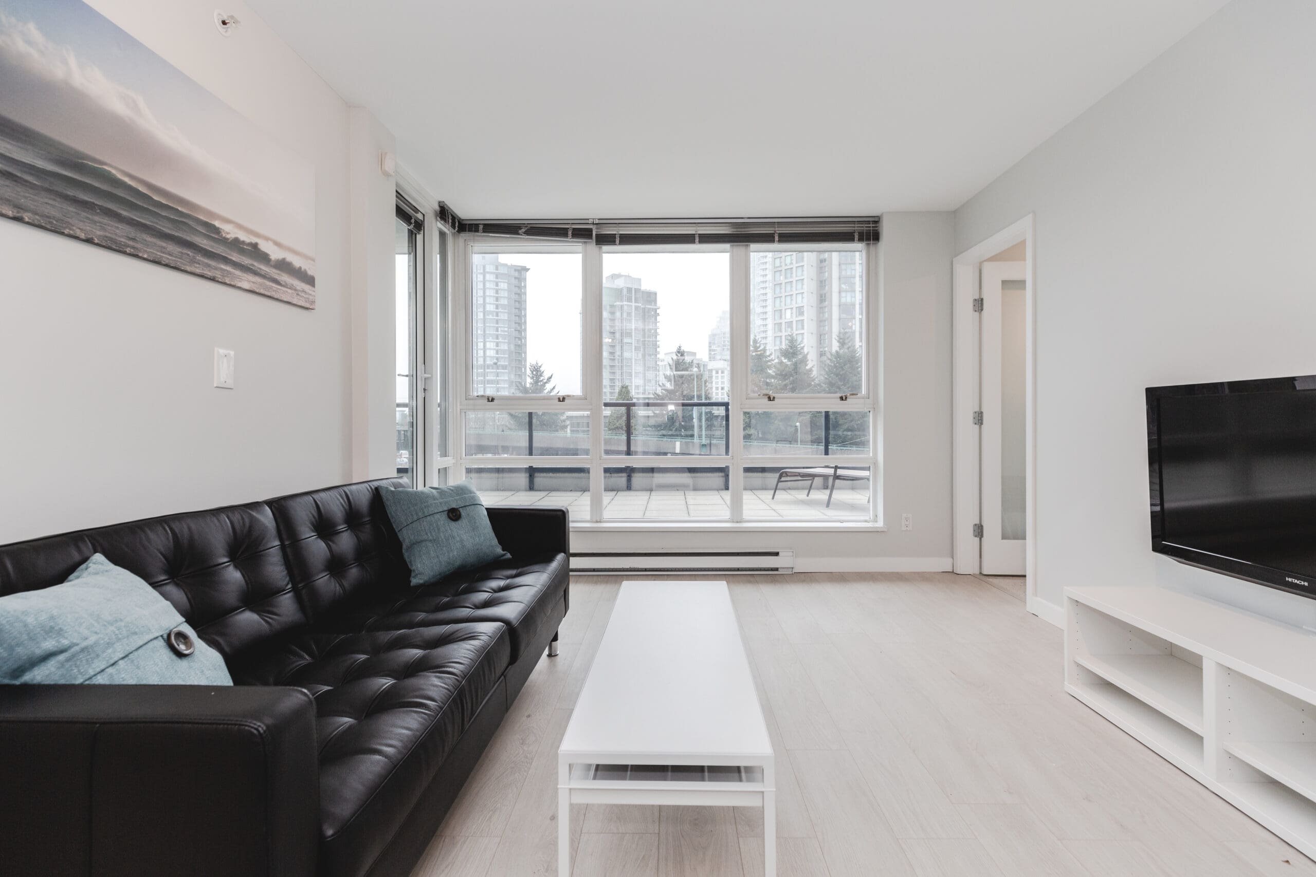 Minimalist living room with a black sofa, white coffee table, and a TV. Large windows offer a view of city buildings.