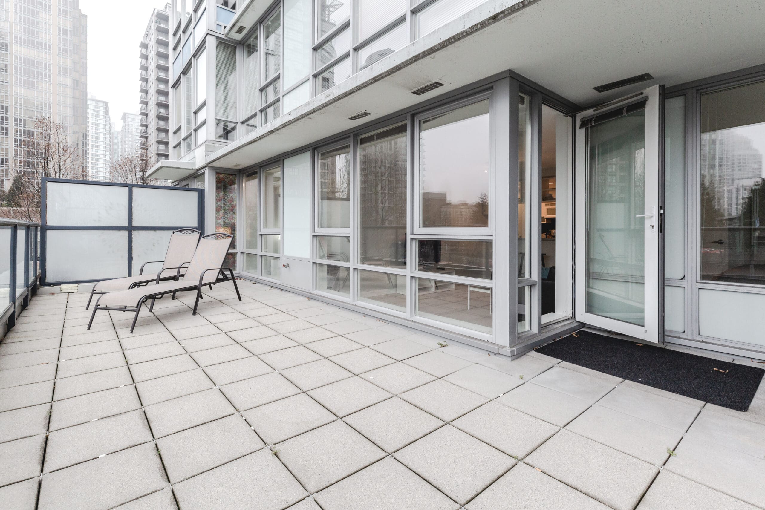 A spacious tiled balcony with two lounge chairs is adjacent to a modern building featuring large glass windows and doors. Surrounding the building are high-rise structures.