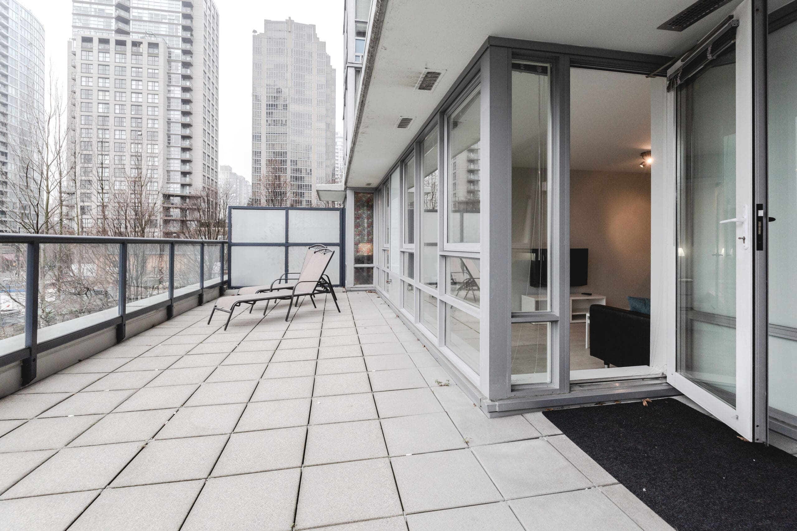 Spacious balcony with two lounge chairs overlooking a cityscape. Glass doors open to a modern living space inside. Urban buildings visible under a cloudy sky.