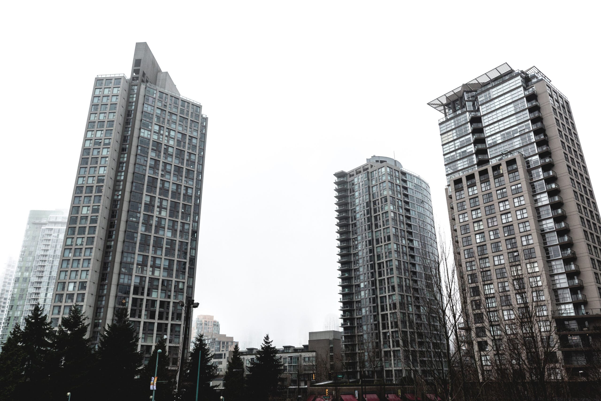 Three tall modern apartment buildings stand against a cloudy sky, surrounded by trees and a few smaller structures.