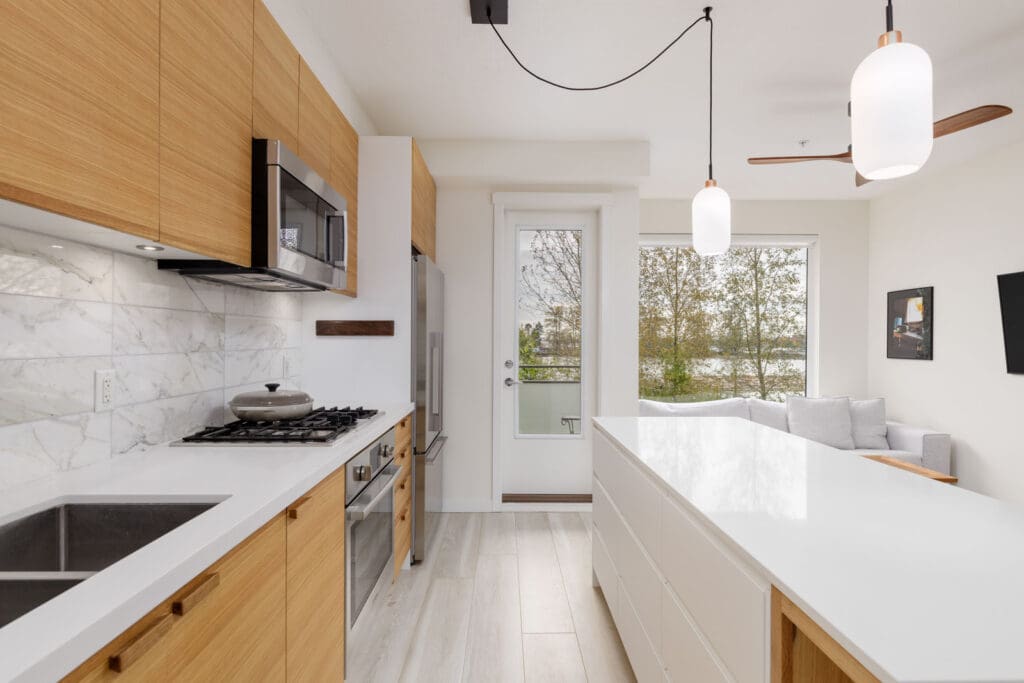 Modern kitchen with wood cabinets, white countertops, and a marble backsplash. Equipped with a gas stove, microwave, and hanging lights. Large window and door to the right.