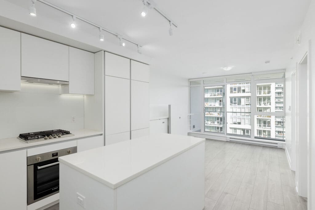 Modern kitchen with white cabinets, island, and built-in stove, adjacent to a bright living area with large windows overlooking a cityscape.