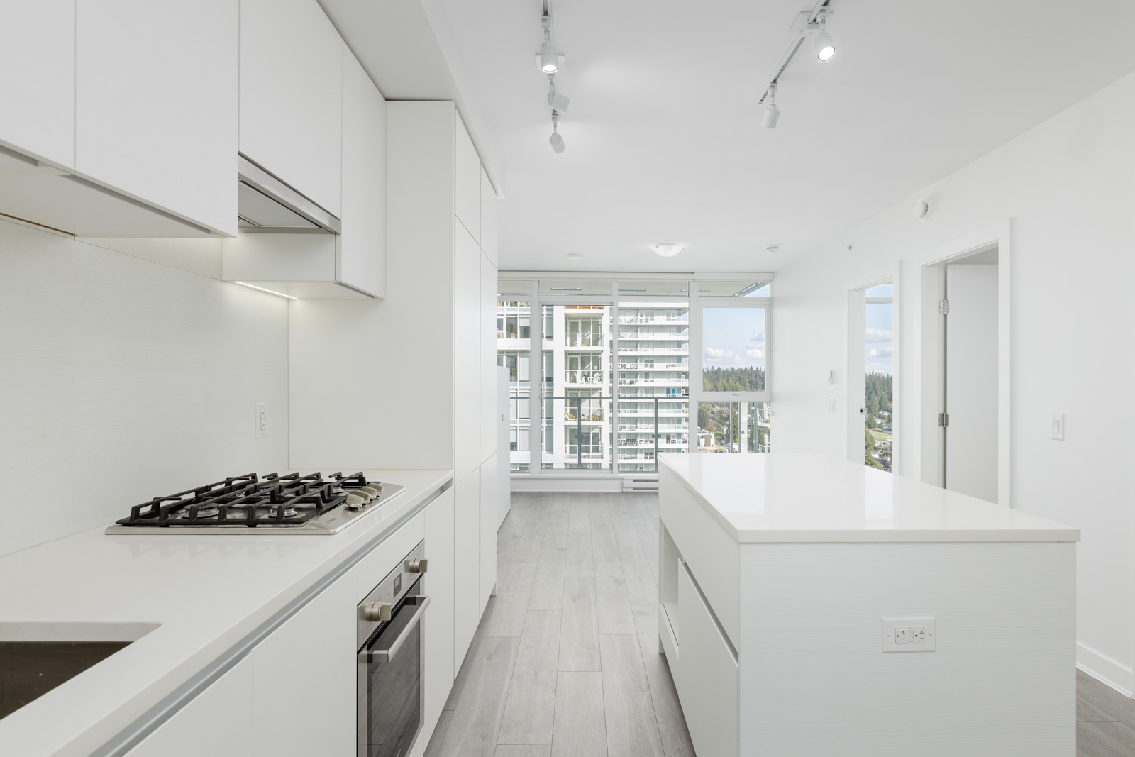 Modern white kitchen with sleek cabinets, a gas stovetop, and an island. Large windows in the background reveal a view of nearby buildings and trees.