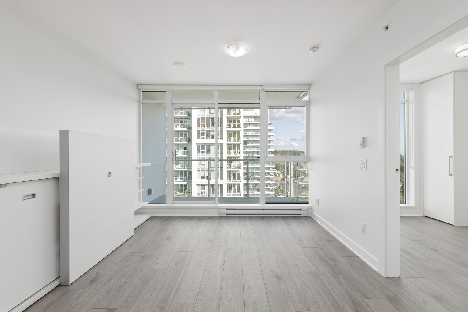 A modern, empty room with light wood flooring, large windows, and a balcony view of buildings and the sky. A fold-down table is on the left wall.