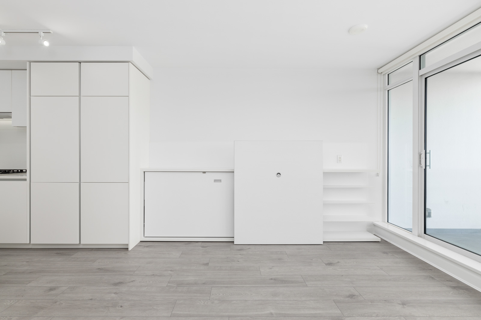 Modern, minimalistic white interior with sleek cabinets, light wood flooring, and large floor-to-ceiling windows on the right.