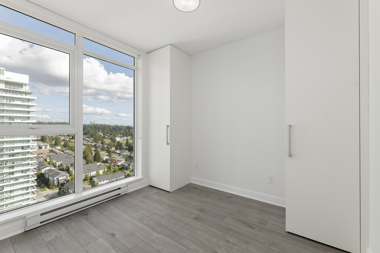 Empty room with light gray flooring, white walls, and a large window offering a cityscape view. There are two tall white cabinets on the right side.