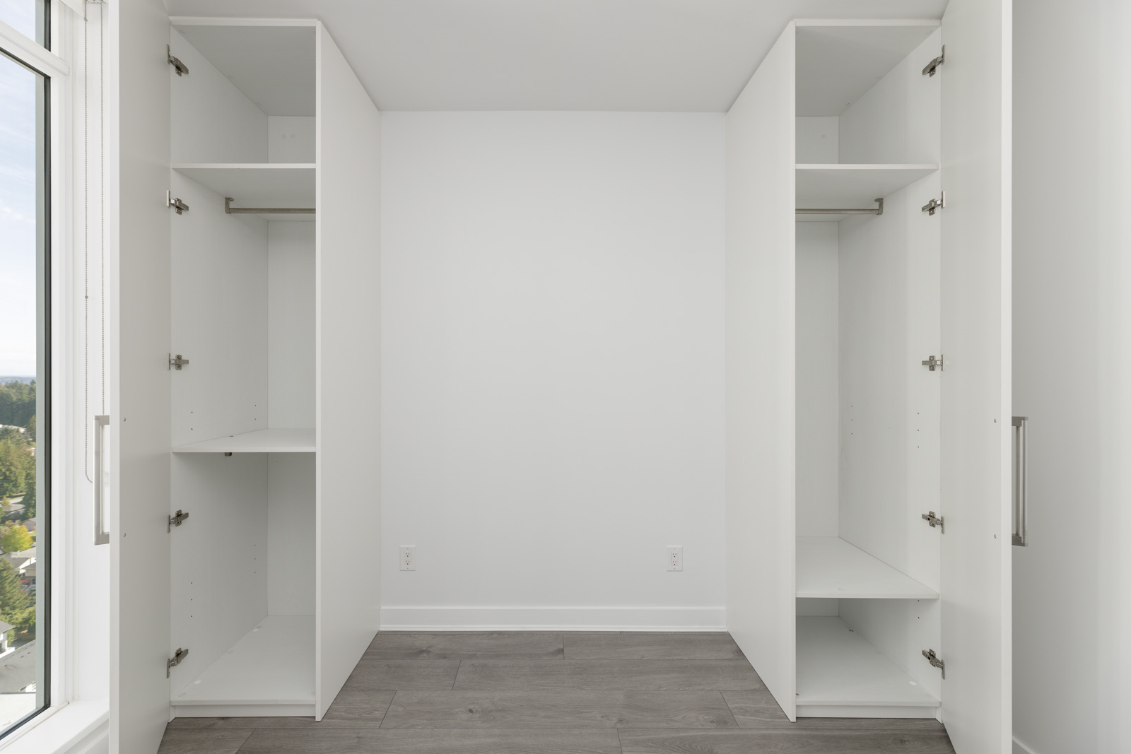 Empty white closet with open doors, featuring shelves and a hanging rod, in a room with gray wood flooring and a window on the left.