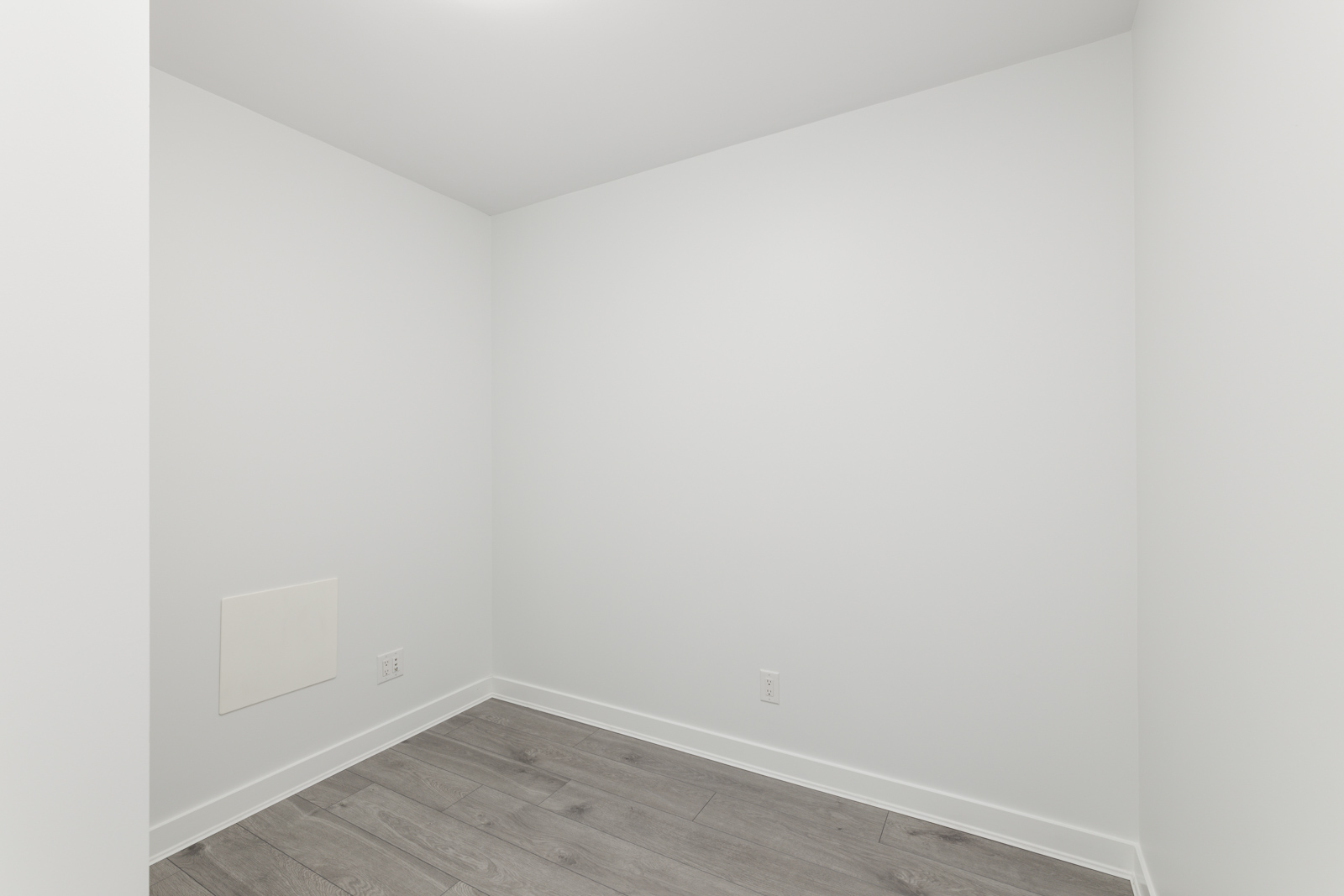 Small empty room with white walls and gray wooden flooring. Electrical outlets are visible on the walls.