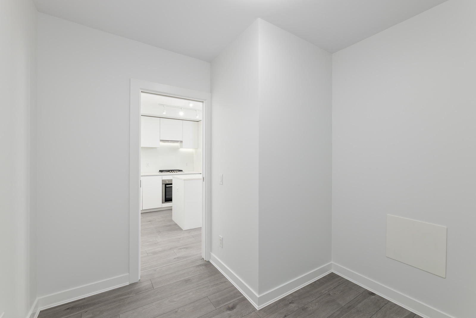 Minimalist room with light gray walls and wood flooring, featuring a doorway leading to a modern kitchen area with white cabinetry and appliances.