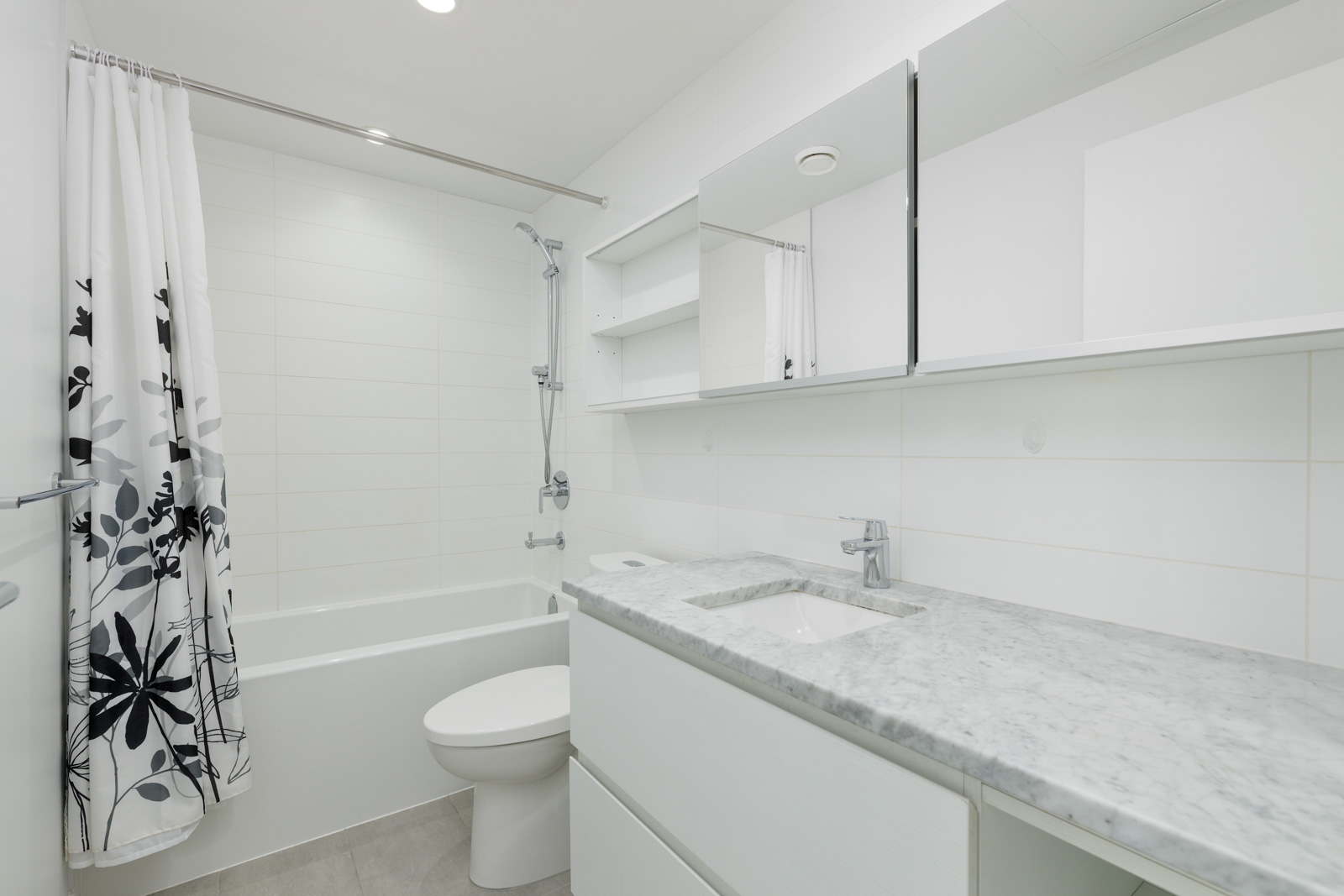 Modern bathroom with a white bathtub, floral shower curtain, marble countertop, sink, toilet, and mirrored cabinet.