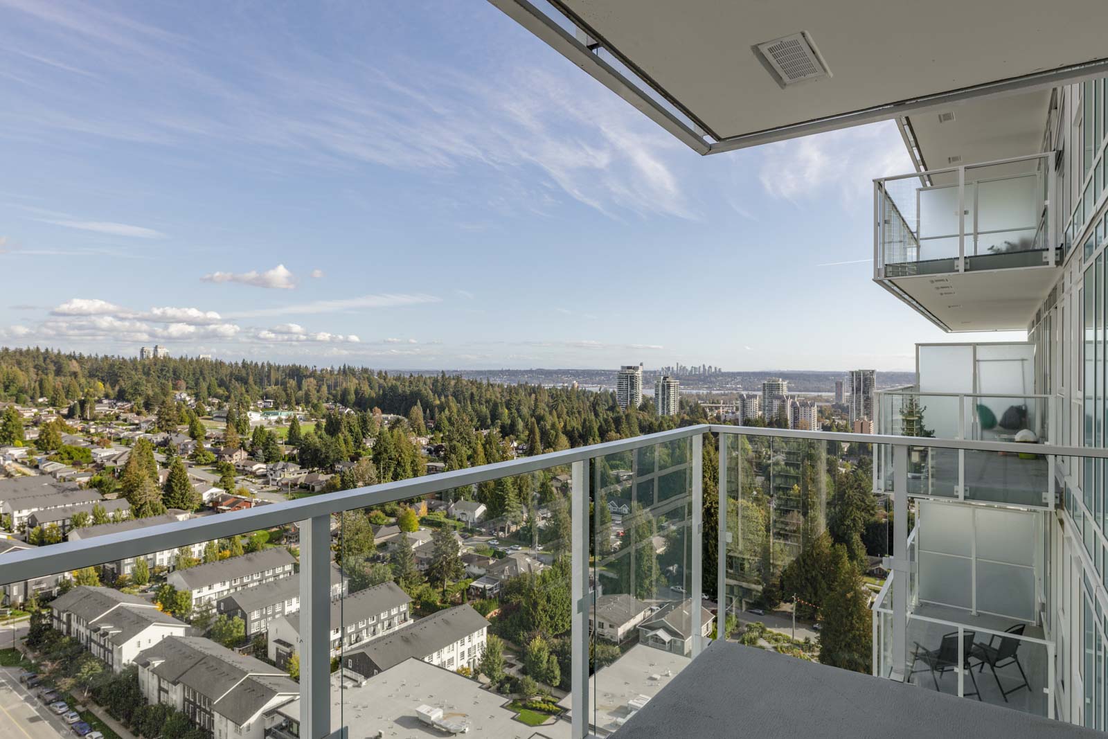View of a cityscape from a high-rise balcony, featuring glass railings, with clear skies and distant buildings and trees.