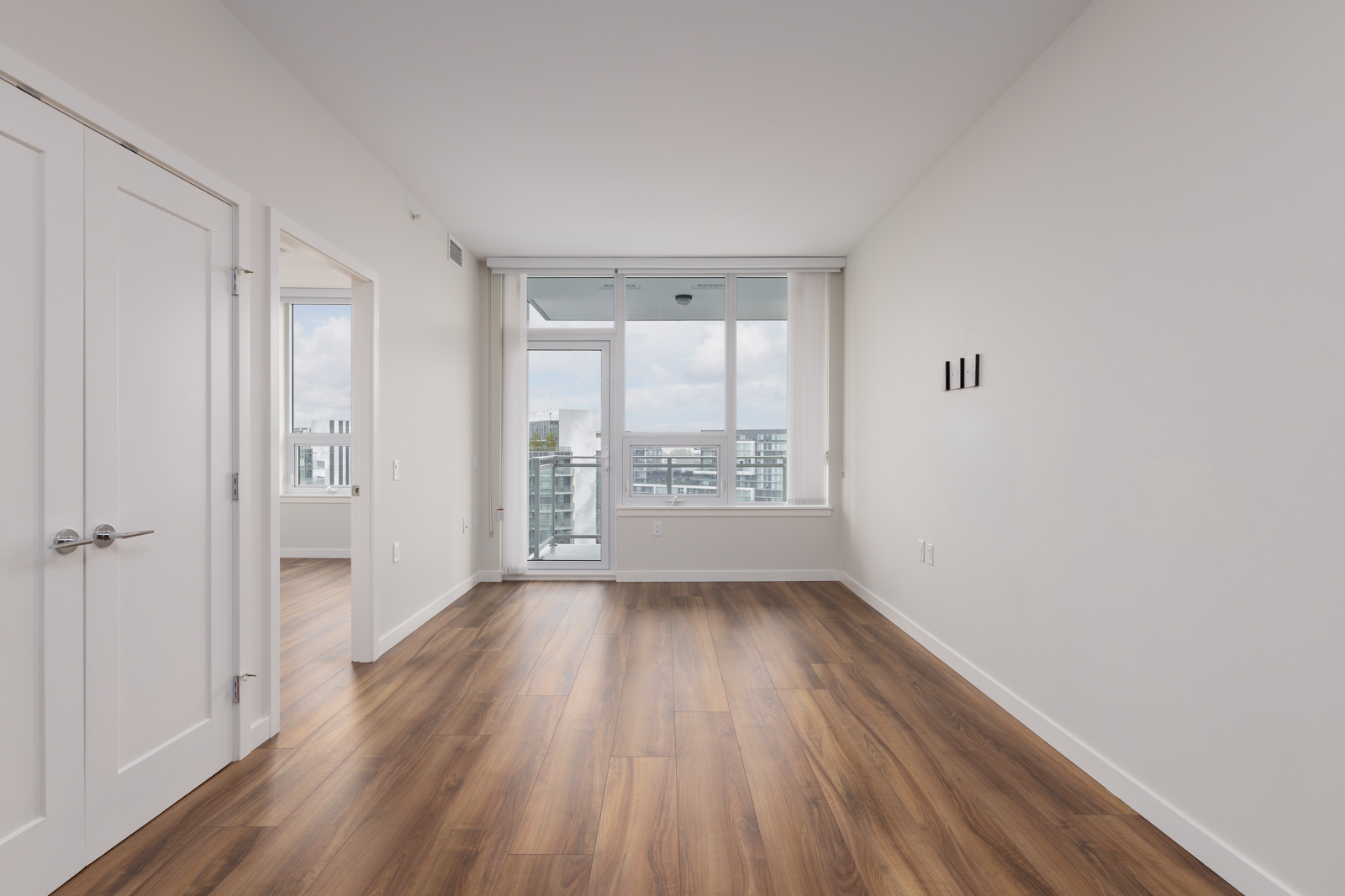 Empty room with wooden floors, a large window with a city view, and white walls. There's a closed double door on the left side.