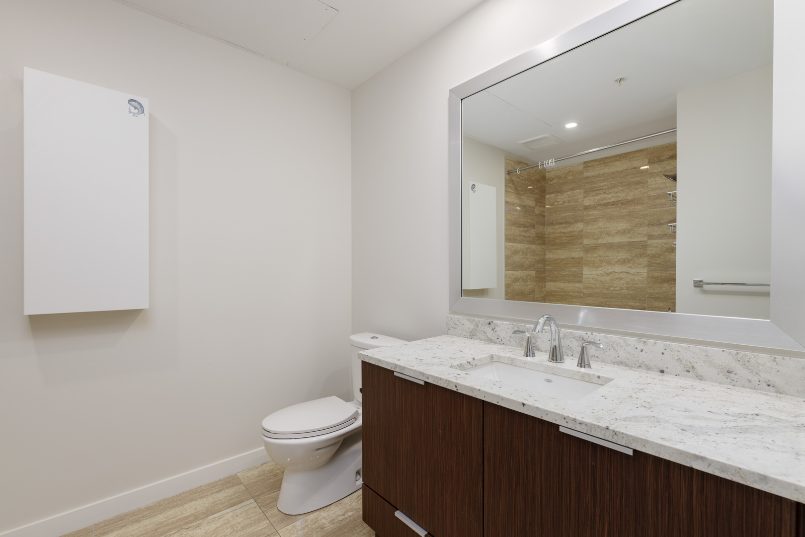Modern bathroom with light wood floors, a white toilet, large wall mirror, and dark wood vanity with double sinks. The shower area features beige tiles.