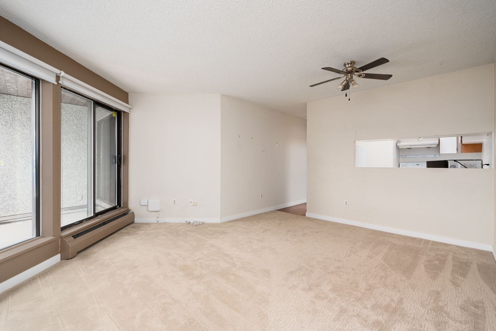 Empty living room with beige carpet, large windows, and a ceiling fan. There's a wall opening to a small kitchen area with visible cabinets and appliances.