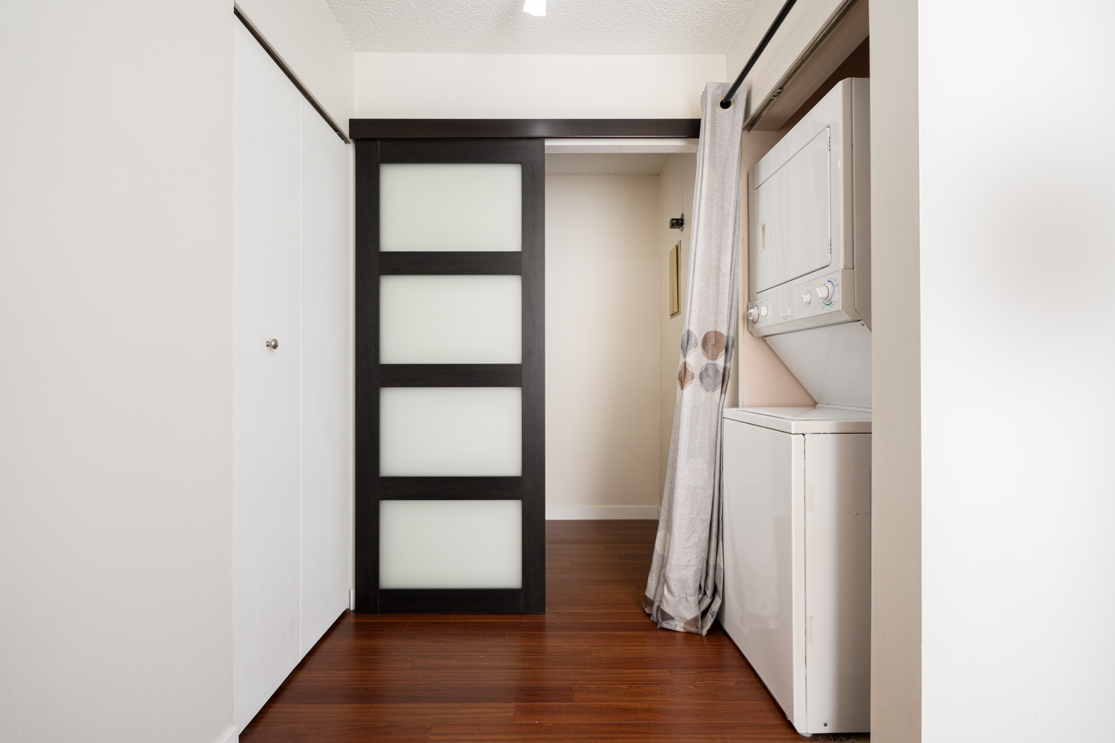 Laundry area with a white stacked washer and dryer to the right, partially hidden by a curtain. A sliding door with frosted glass panels is on the left. The floor is wooden.