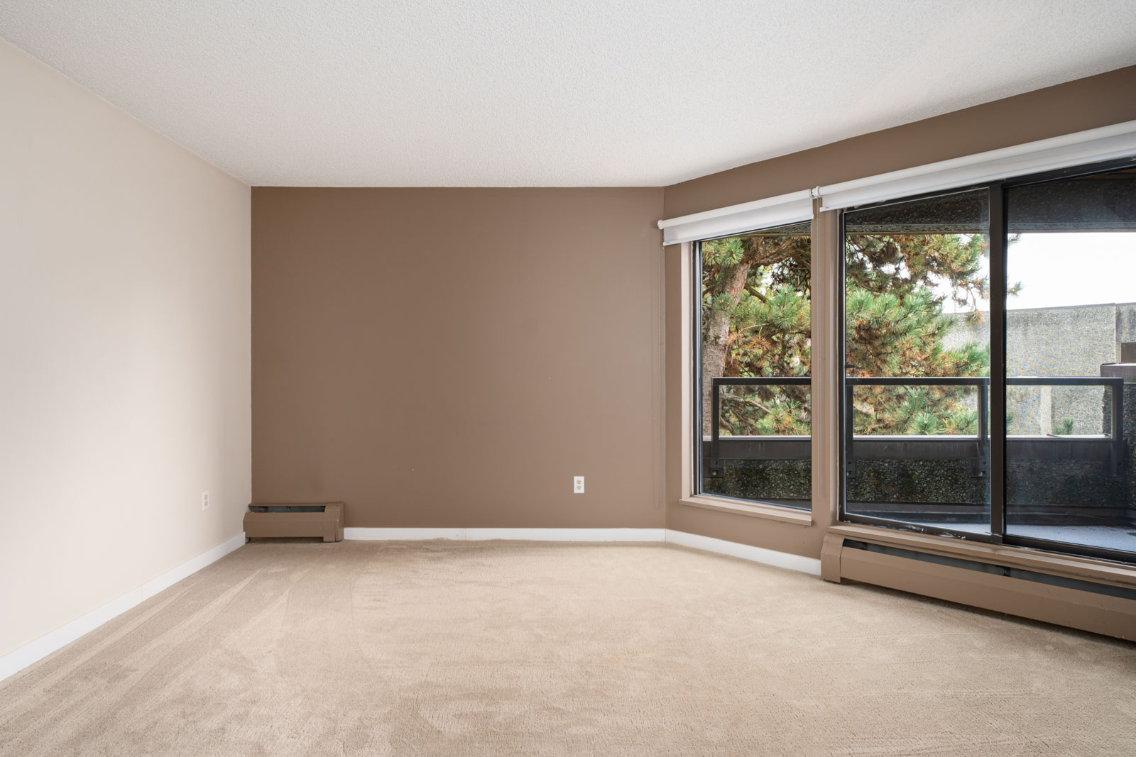 Empty room with beige carpet, brown accent wall, and large windows overlooking a balcony with trees outside.