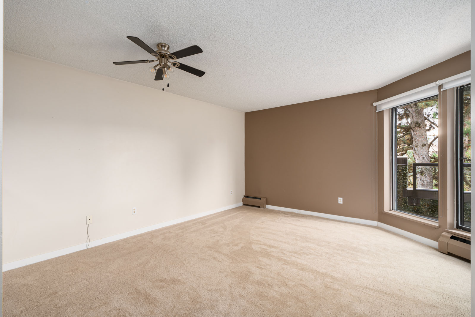 A vacant carpeted room with beige walls, a ceiling fan, and a large window with white blinds.