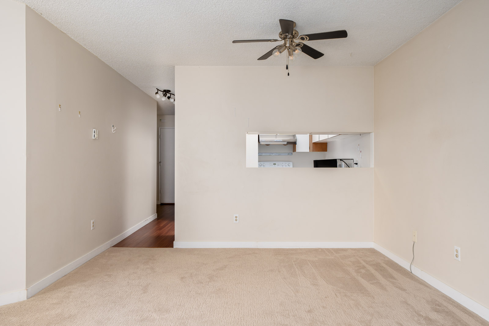 An empty room with beige walls and carpet, a ceiling fan, and a wall cut-out revealing a kitchen area with white appliances and hardwood flooring.