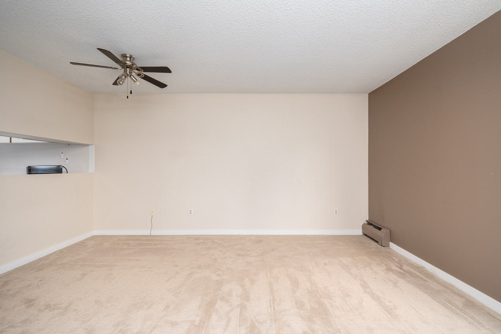 Empty room with beige carpet, two-tone walls, a ceiling fan, and a small open kitchen space in the corner.