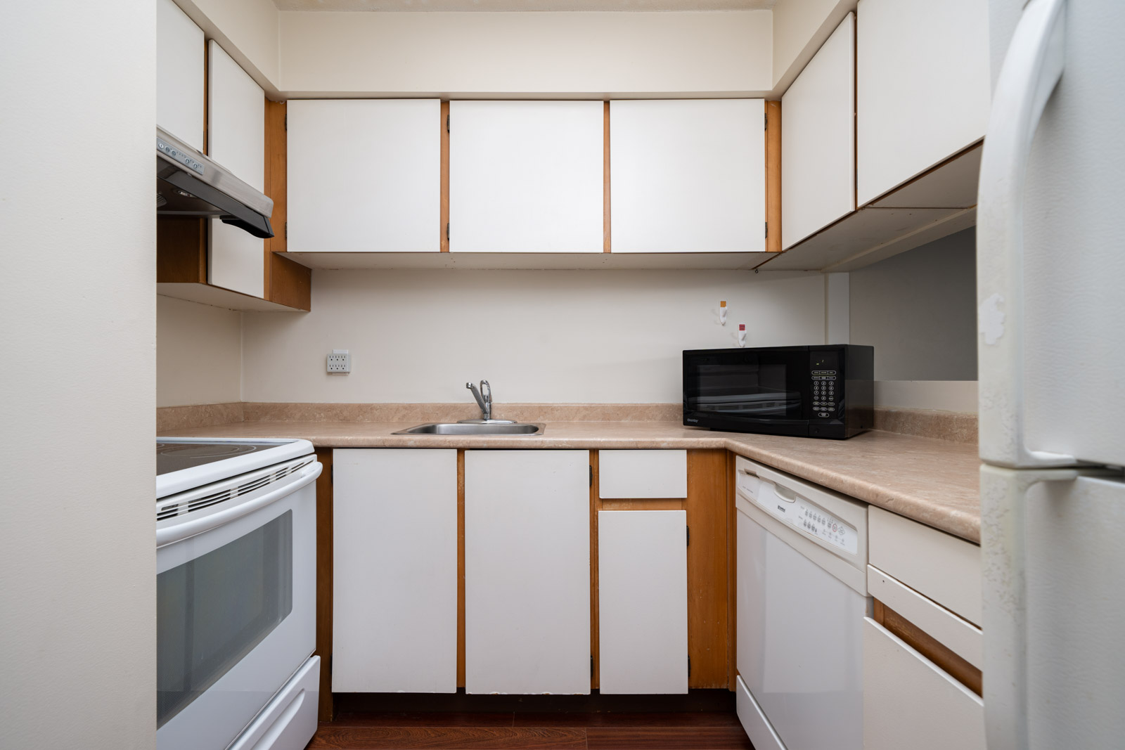 Compact kitchen with white cabinets, an oven, a microwave, a dishwasher, and a fridge. Brown countertops and a stainless steel sink are visible.
