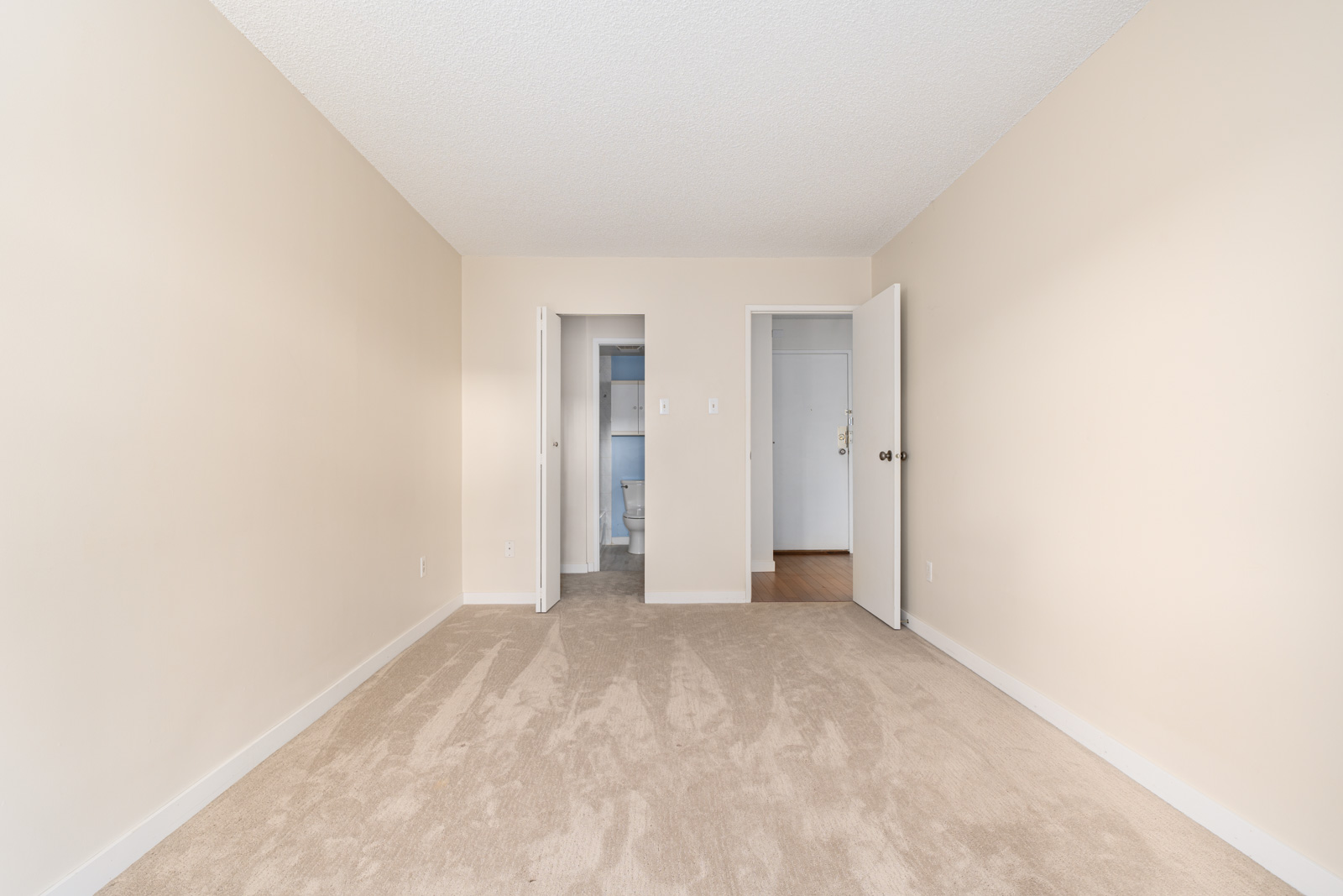 Empty beige carpeted room with an open door leading to a closet and another open door revealing a bathroom. Walls are plain, and the ceiling is textured.