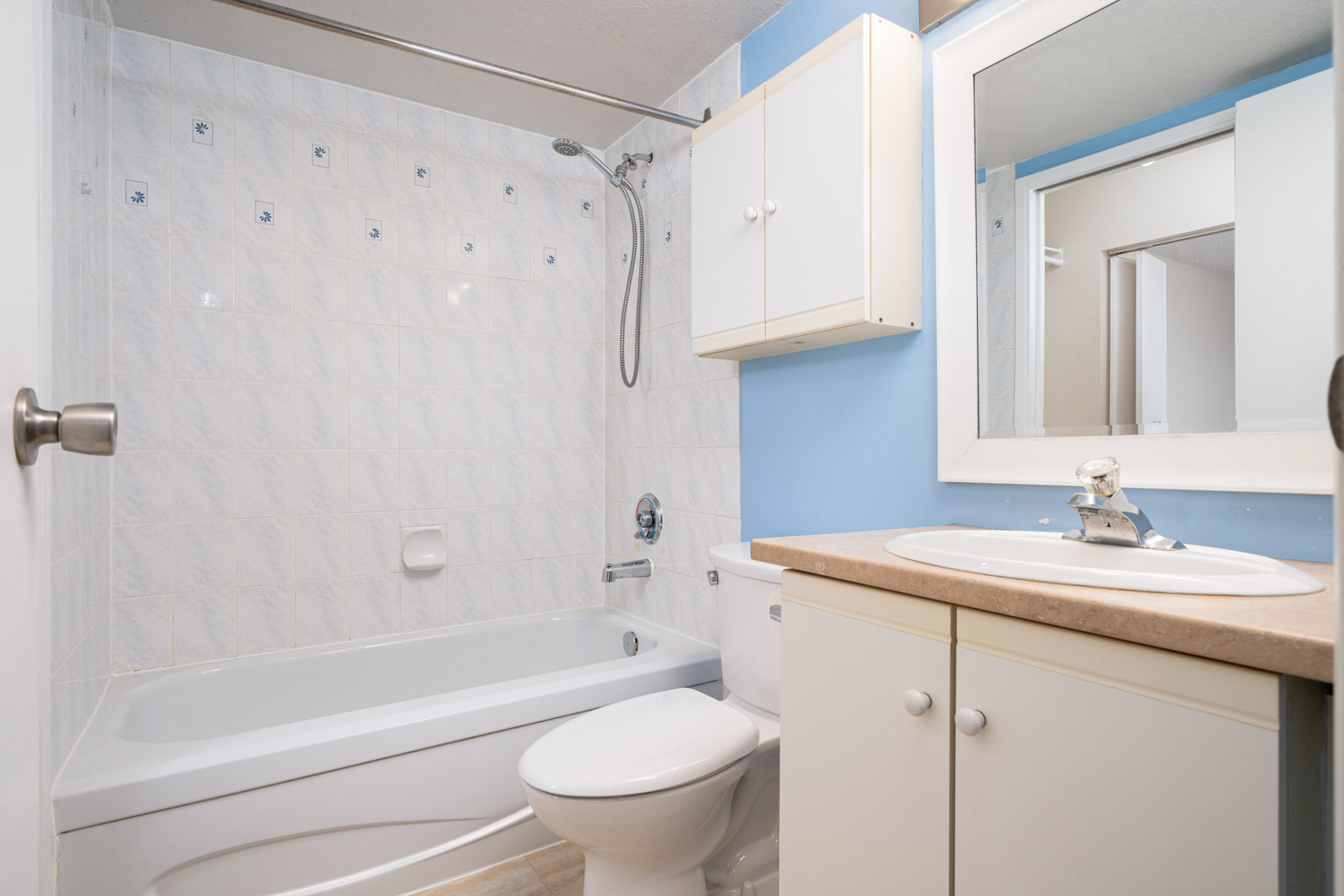 A clean bathroom with a white bathtub, showerhead, toilet, sink, and cabinets. The walls are partially tiled and painted blue, with a mirror above the sink.