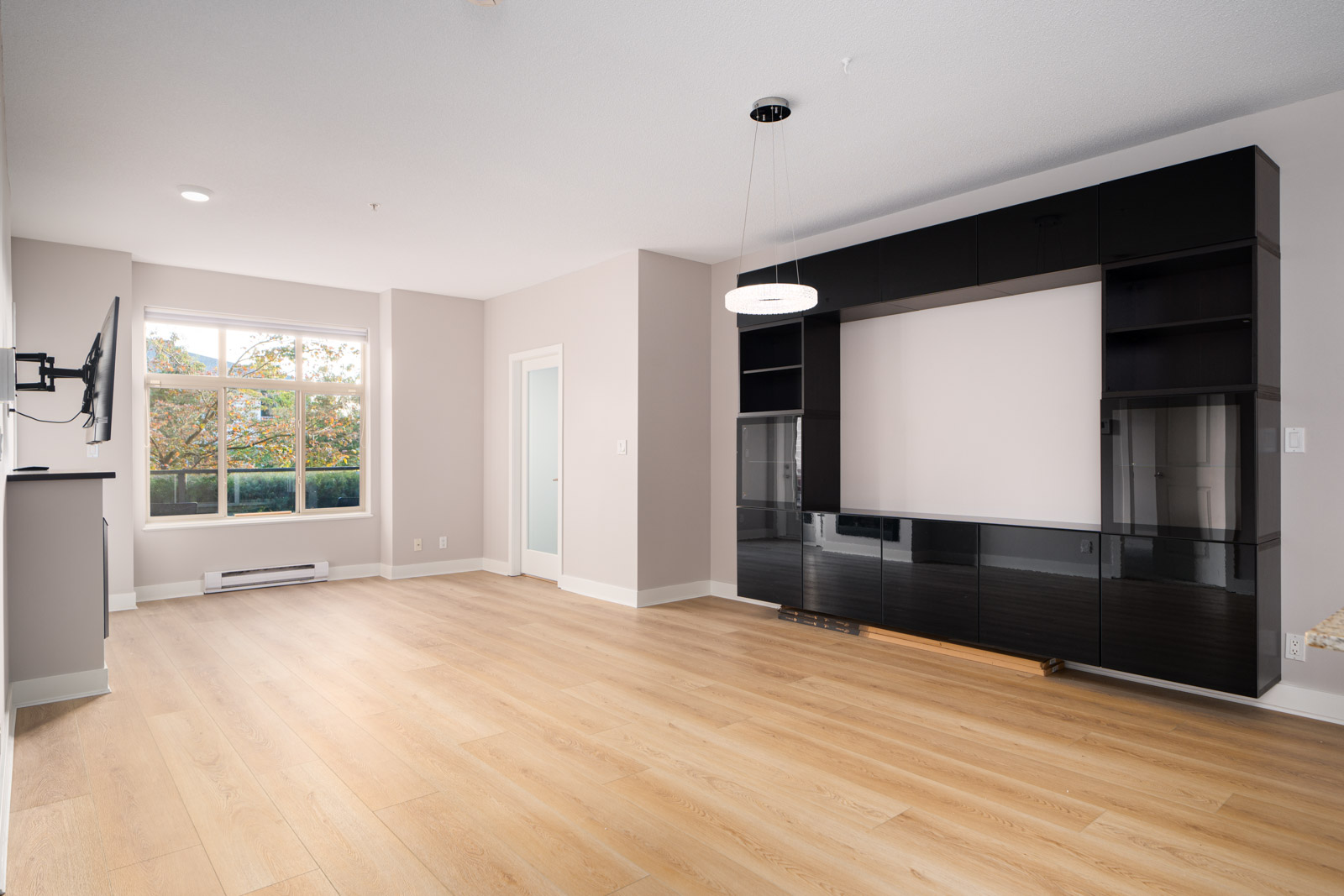 An empty living room with light wood flooring, large window, gray walls, and a black entertainment unit against one wall. A modern ceiling light hangs above.