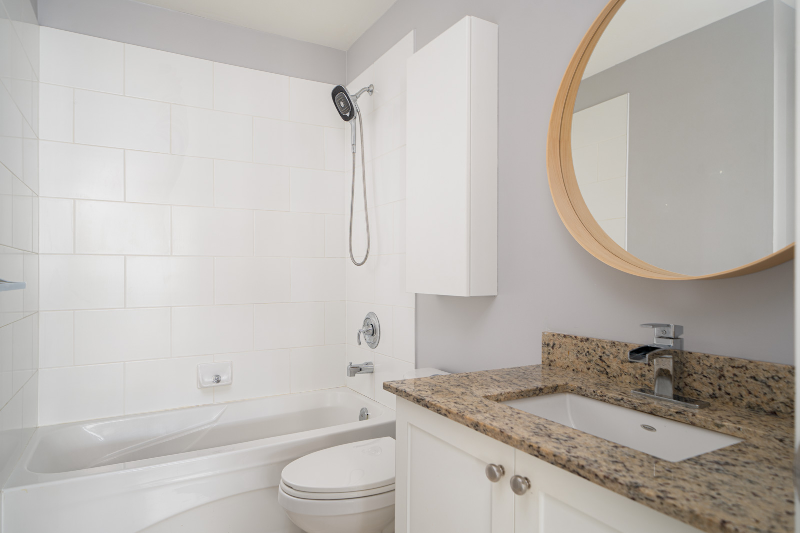 Modern bathroom with white tiles, bathtub, and overhead shower. Granite countertop with sink, faucet, and circular mirror above. White cabinets below the sink.