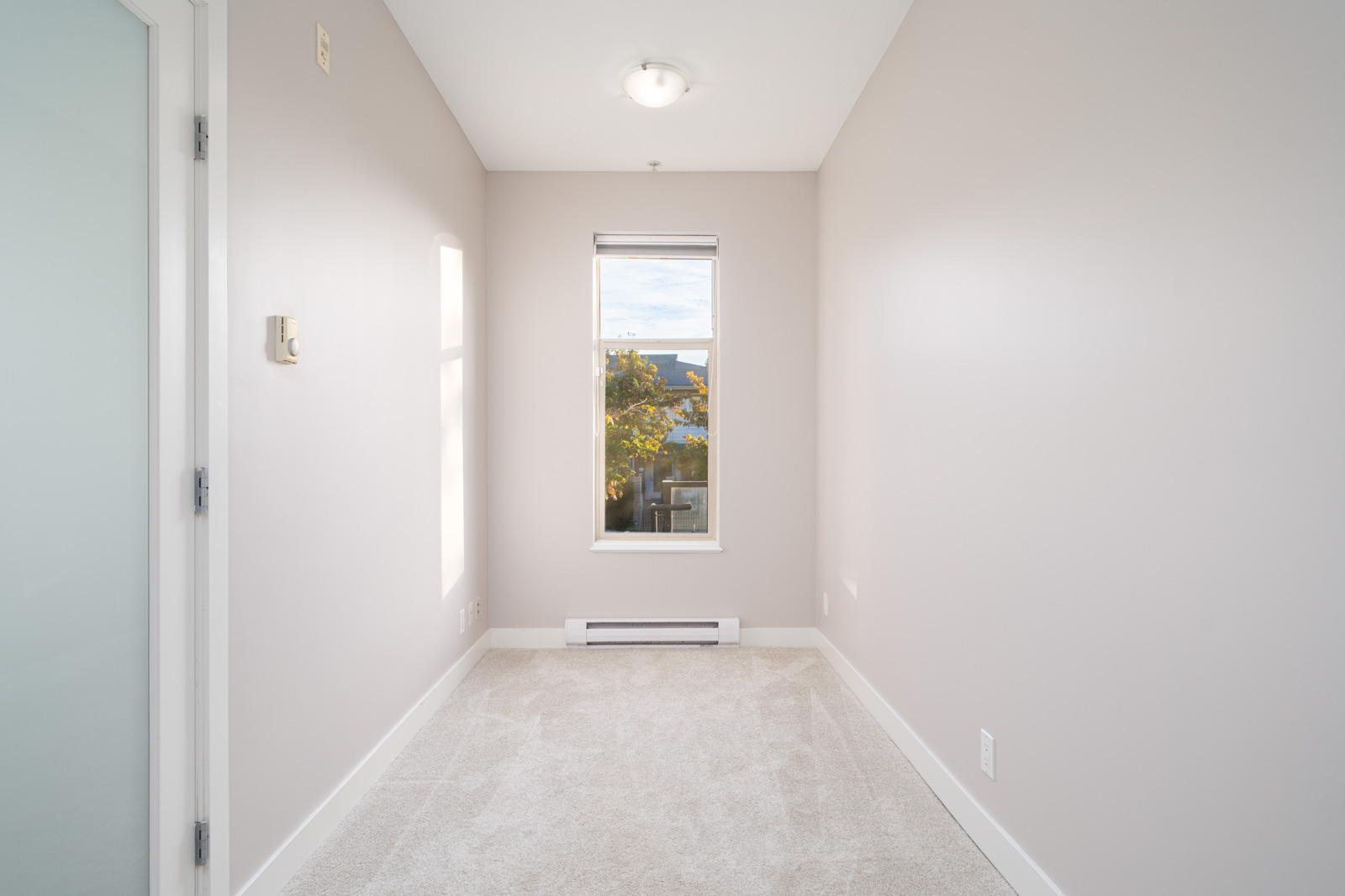 Small, empty room with beige walls, a single window showing trees outside, and a white ceiling light fixture. The floor is carpeted, and a wall heater is below the window.