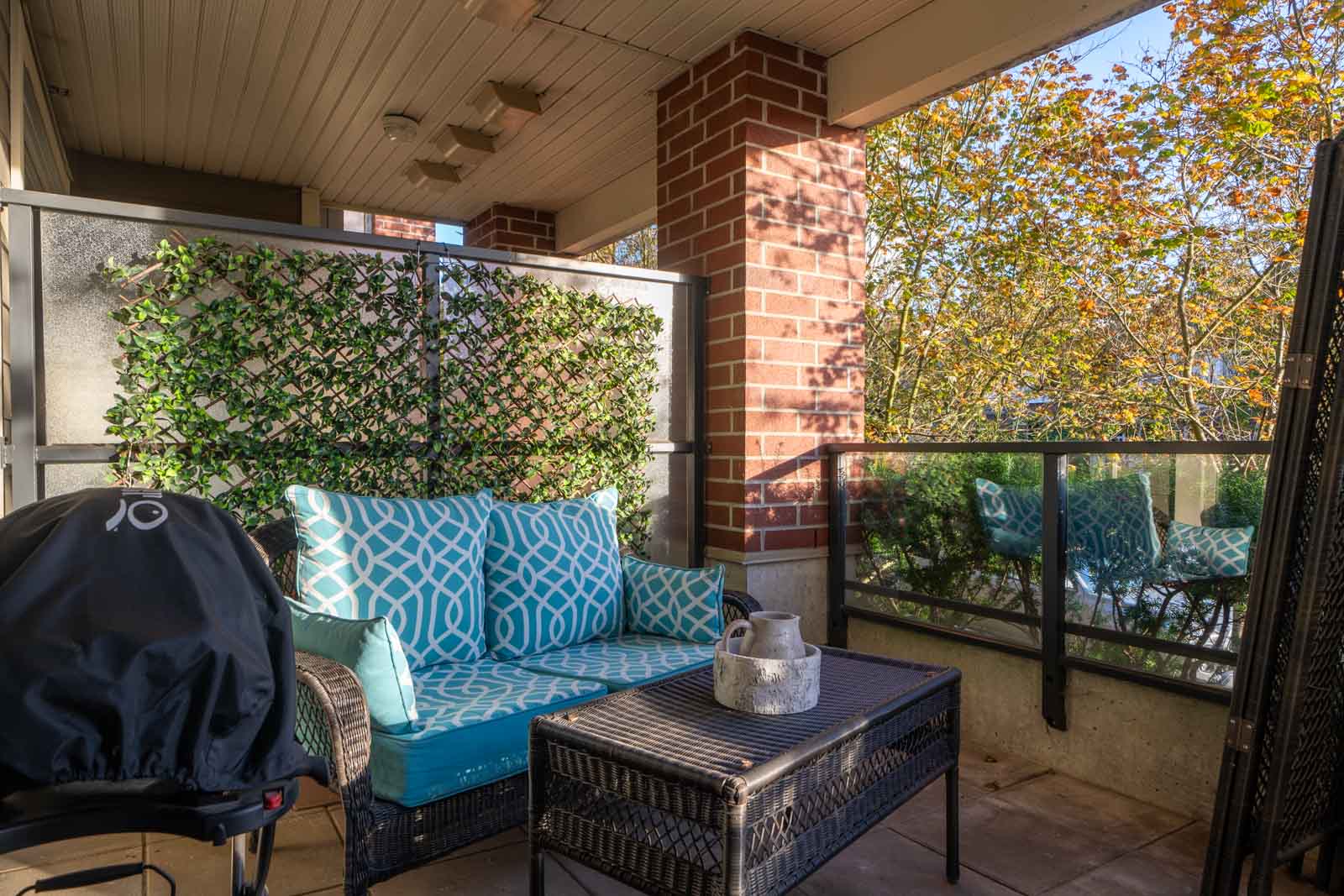 A cozy patio with a blue cushioned loveseat, a black wicker table, a small barbecue grill, and leafy privacy panels. Sunlight filters through autumn trees in the background.