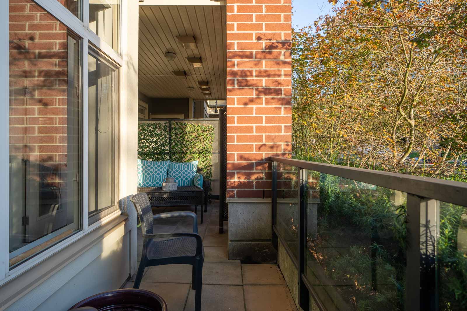 A small balcony with brick walls, glass railing, and two chairs. A tree with autumn leaves is visible outside.
