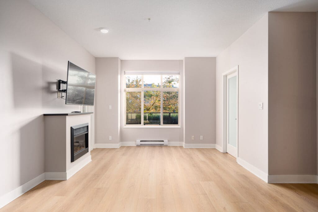 Bright, empty living room with light wood flooring, a wall-mounted TV above a fireplace, and large windows with a view of trees.