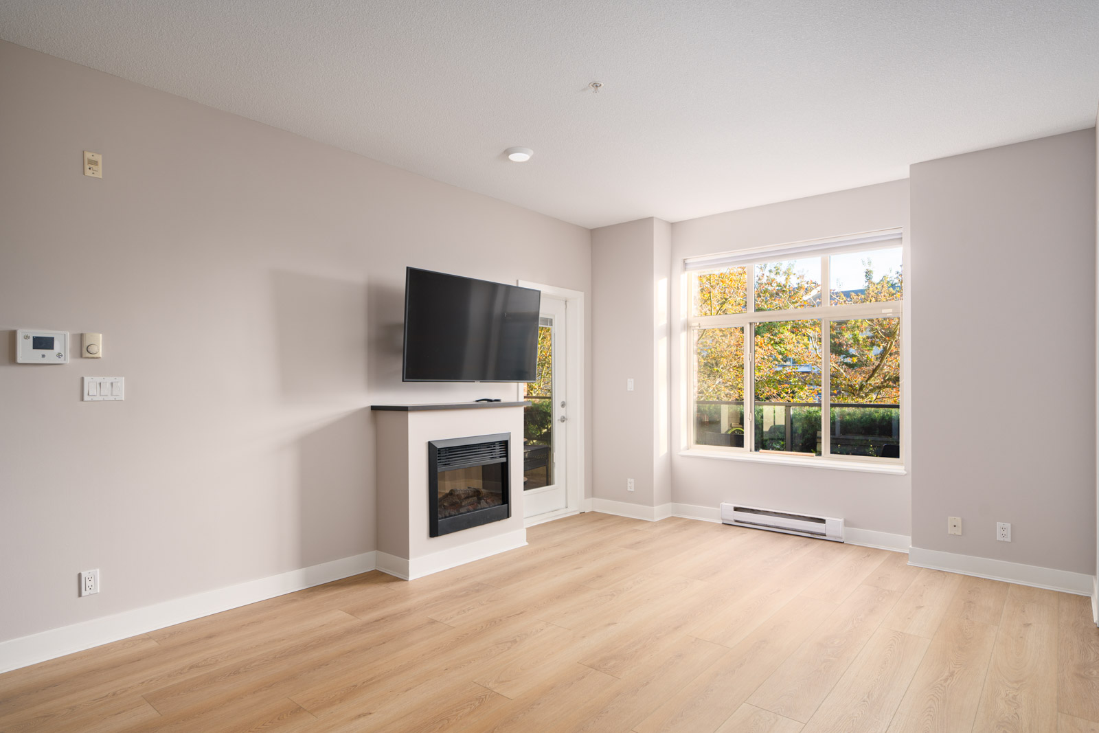 Bright, empty living room with light wood flooring, a wall-mounted TV above a fireplace, and a large window showing greenery outside.