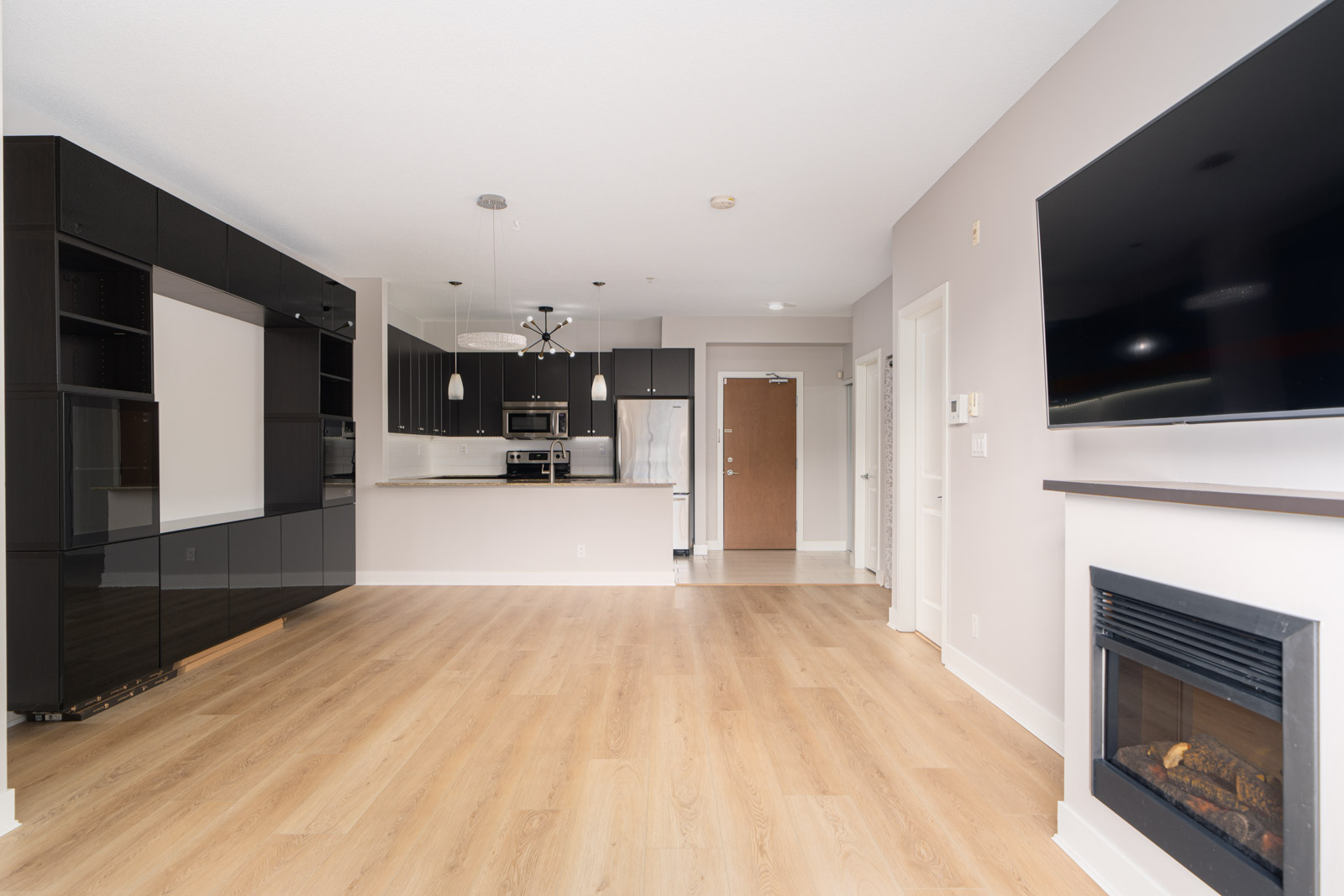 Modern open-concept living area with a kitchen, black cabinets, wood flooring, TV on the wall, and an electric fireplace.