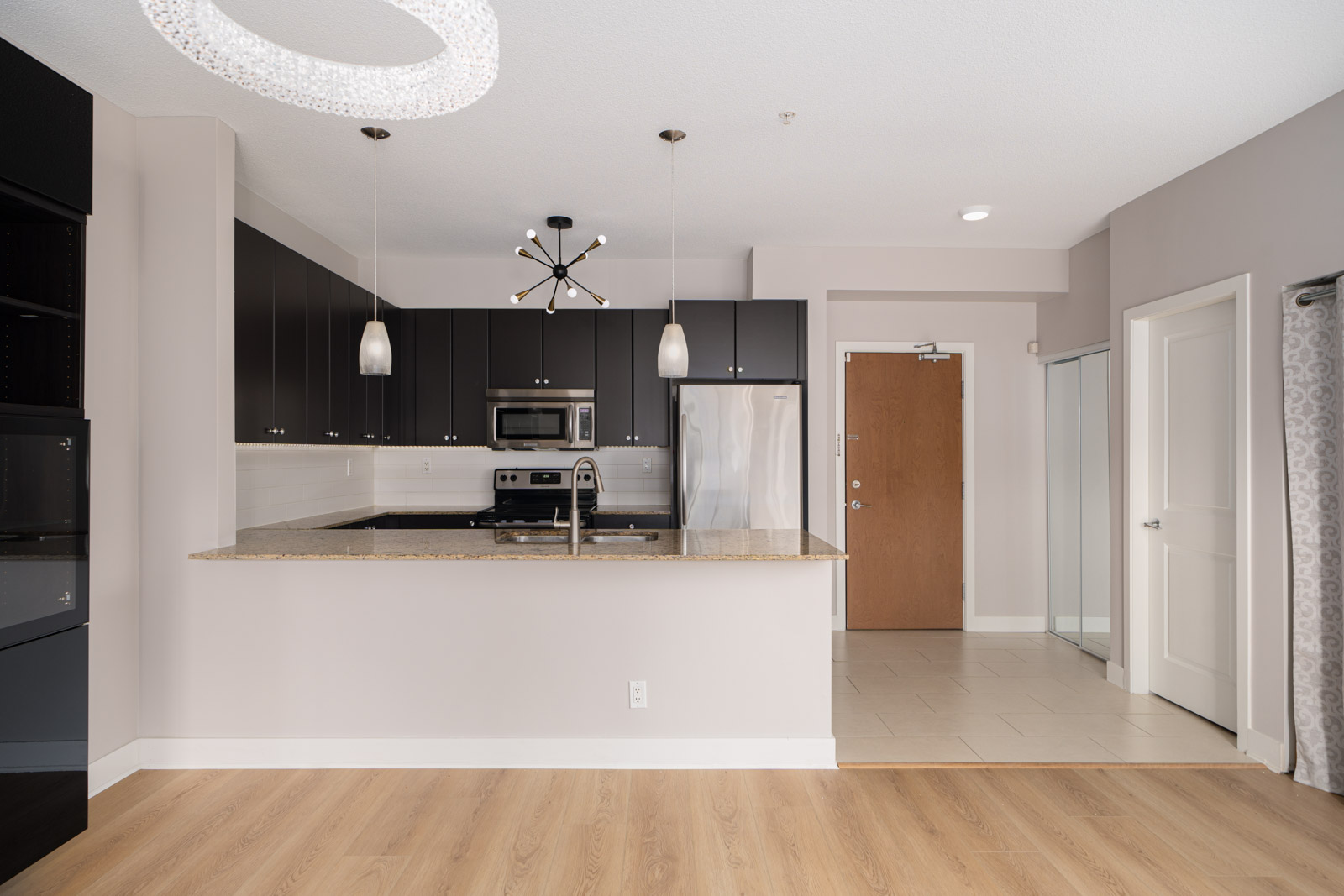 Modern kitchen with black cabinets, stainless steel appliances, and a granite countertop. Pendant lights hang over the island. A wooden floor contrasts with the light walls and ceiling.