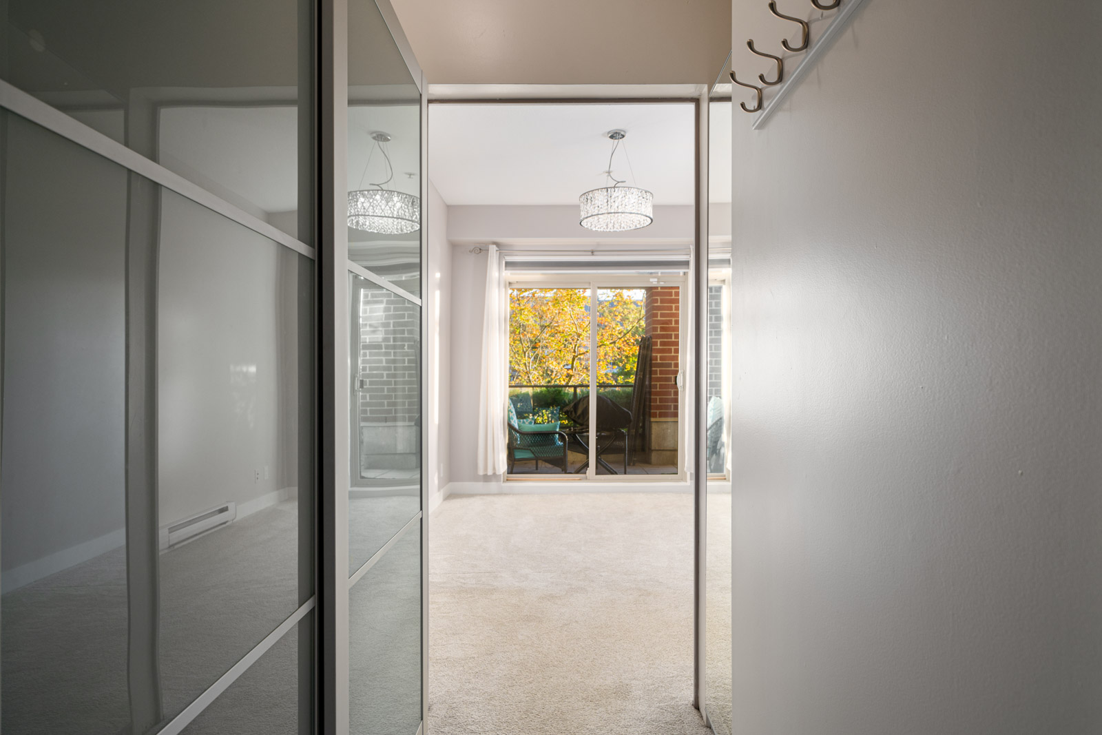 Hallway with mirrored closet doors leads to a carpeted room with a glass door opening to a balcony with trees visible outside. A chandelier is hanging from the ceiling.