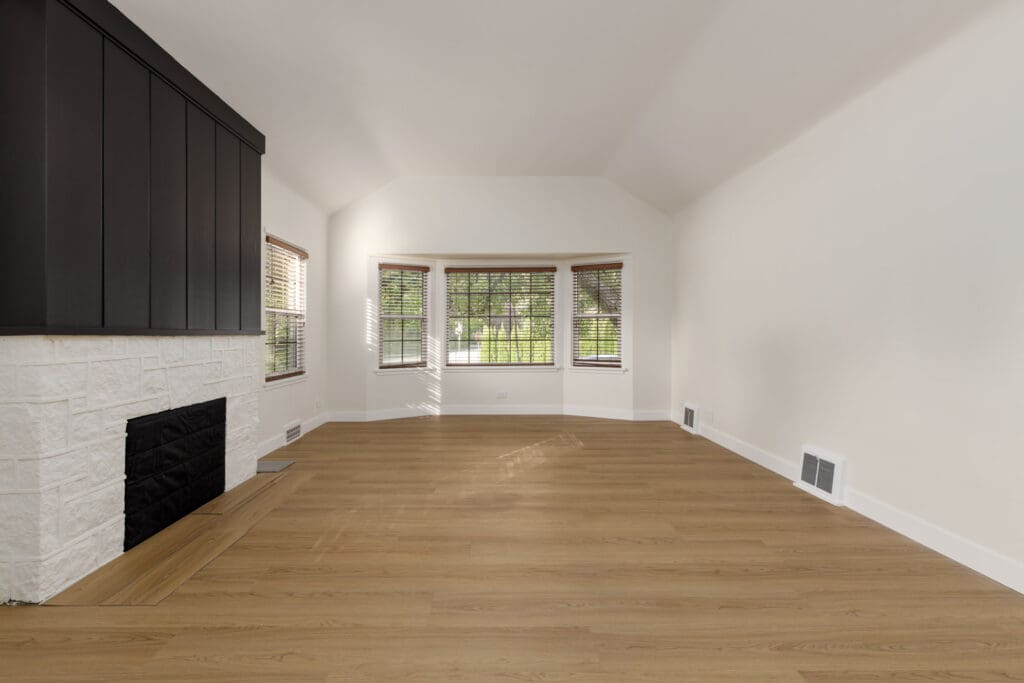 Empty living room with light wood flooring, a black and white fireplace, and a bay window with blinds. Walls and ceiling are painted white.
