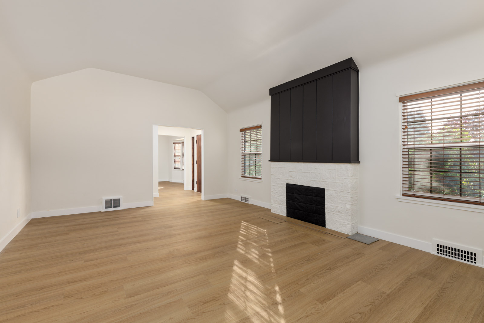 Empty room with light wood flooring, white walls, a white brick fireplace with a dark mantle, and two windows with blinds.