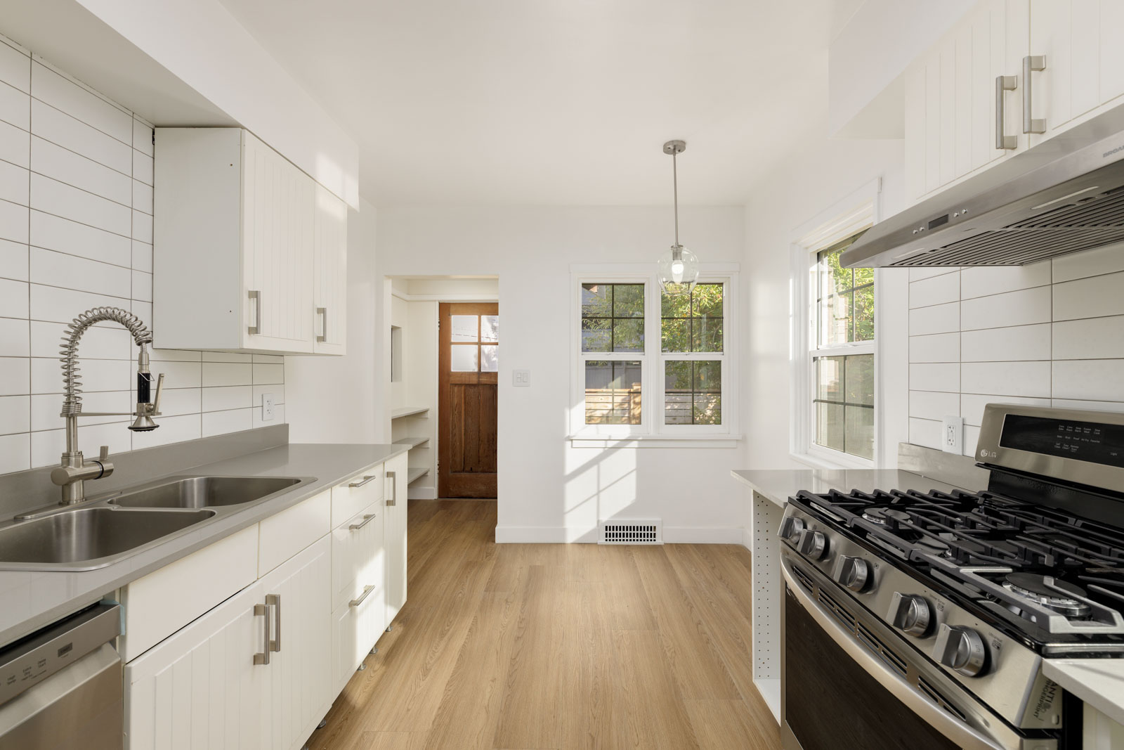 Modern kitchen with white cabinets, stainless steel appliances, gas stove, and wood flooring. Natural light from windows and a door at the far end.