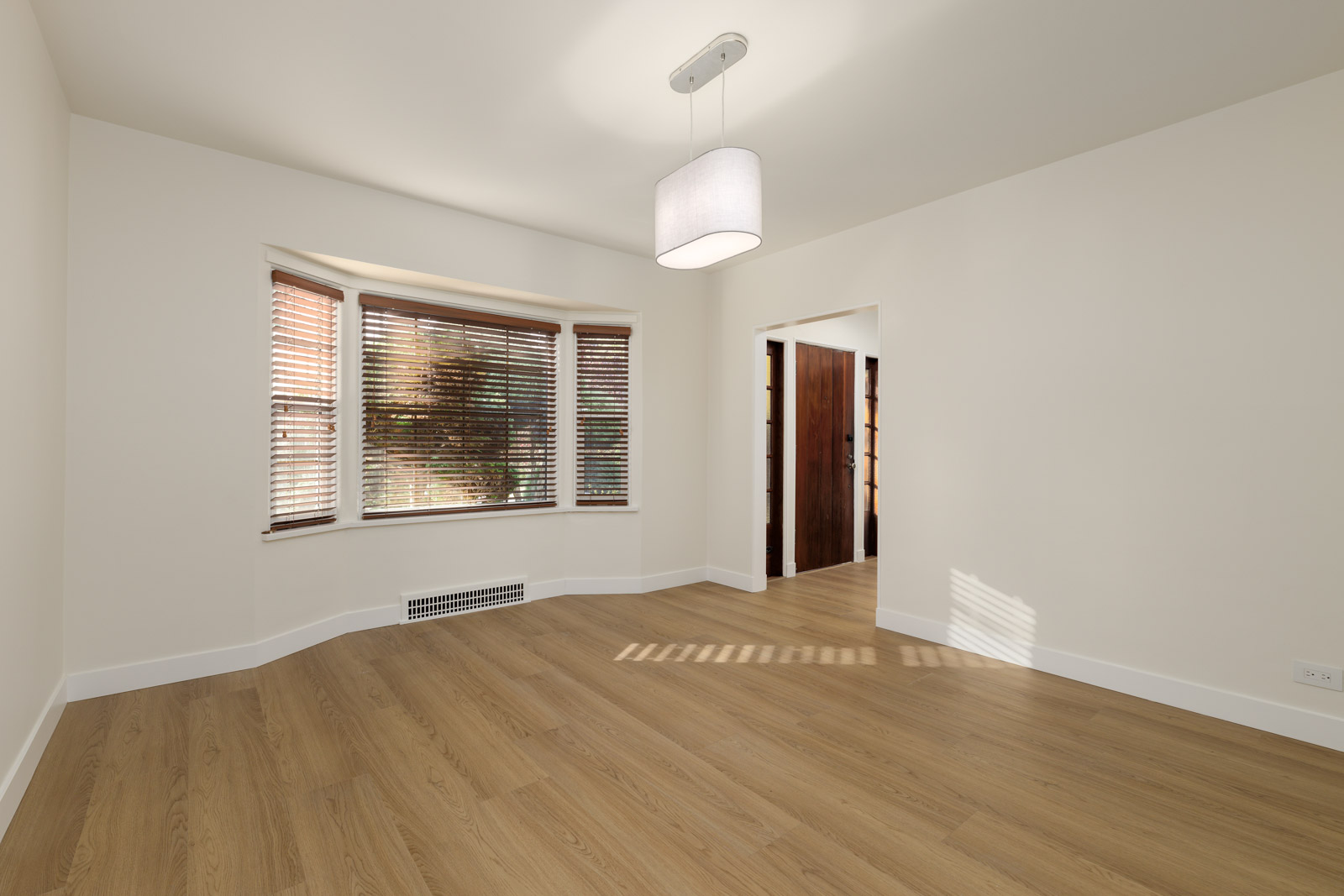 An empty room with light wood flooring, white walls, a window with blinds, and a ceiling light fixture. An open doorway leads to another area.