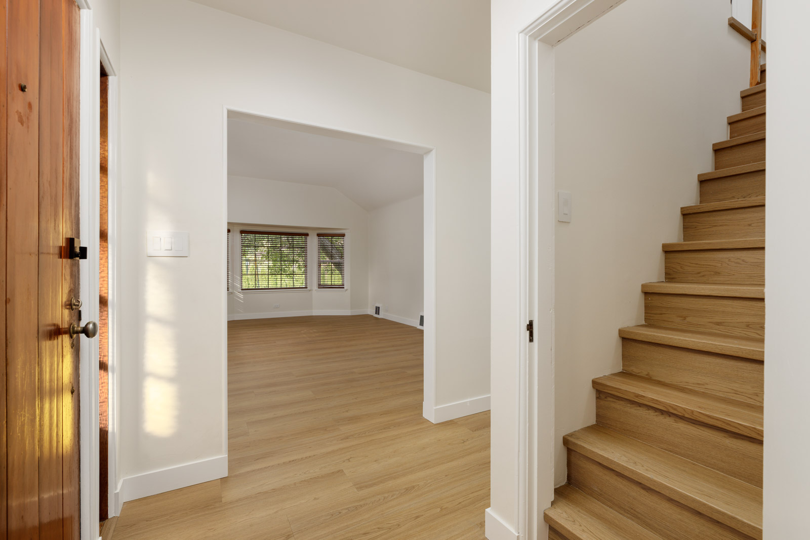 Interior view of an empty room with wooden flooring, open doorway leading to another room with large windows, and a staircase on the right side.