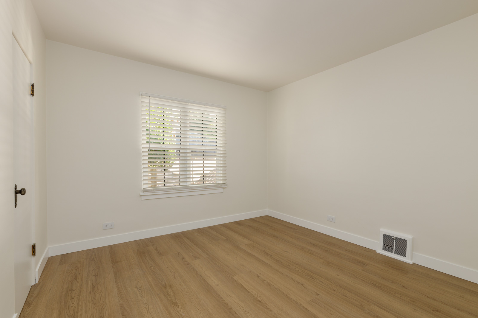 An empty room with light beige walls, wooden flooring, a window with blinds, a white door, and a small wall vent.