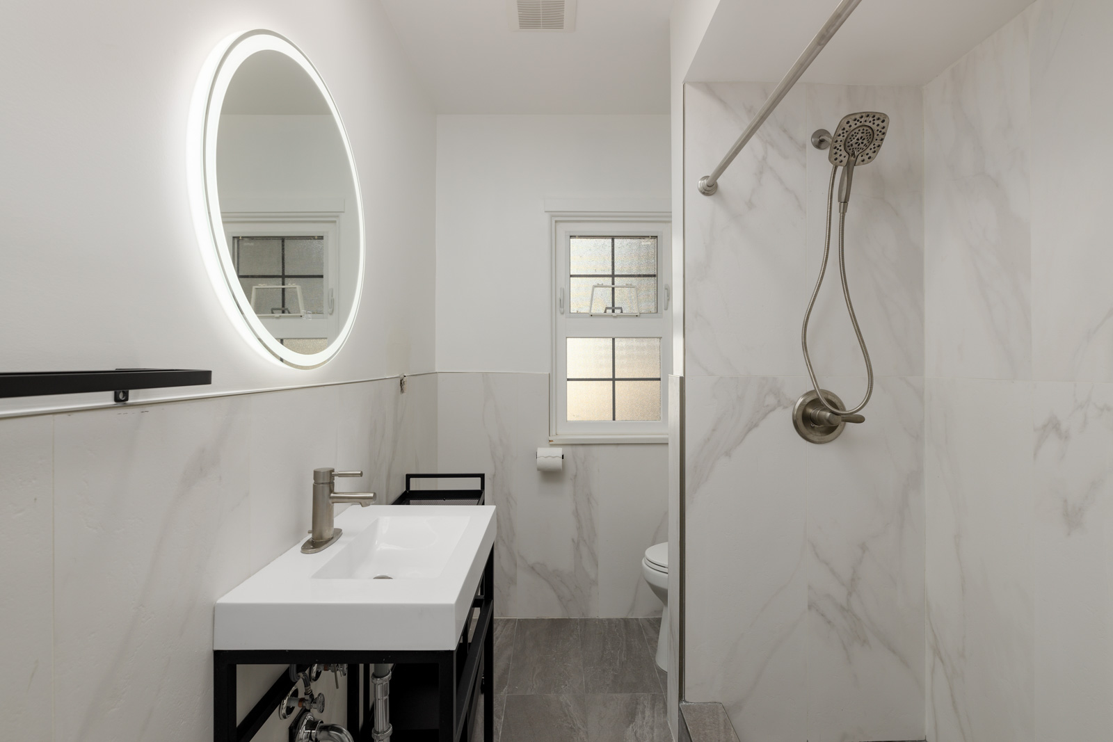 Modern bathroom with a circular illuminated mirror, rectangular sink, and wall-mounted showerhead. White walls and tiled surfaces create a clean and minimalist look.